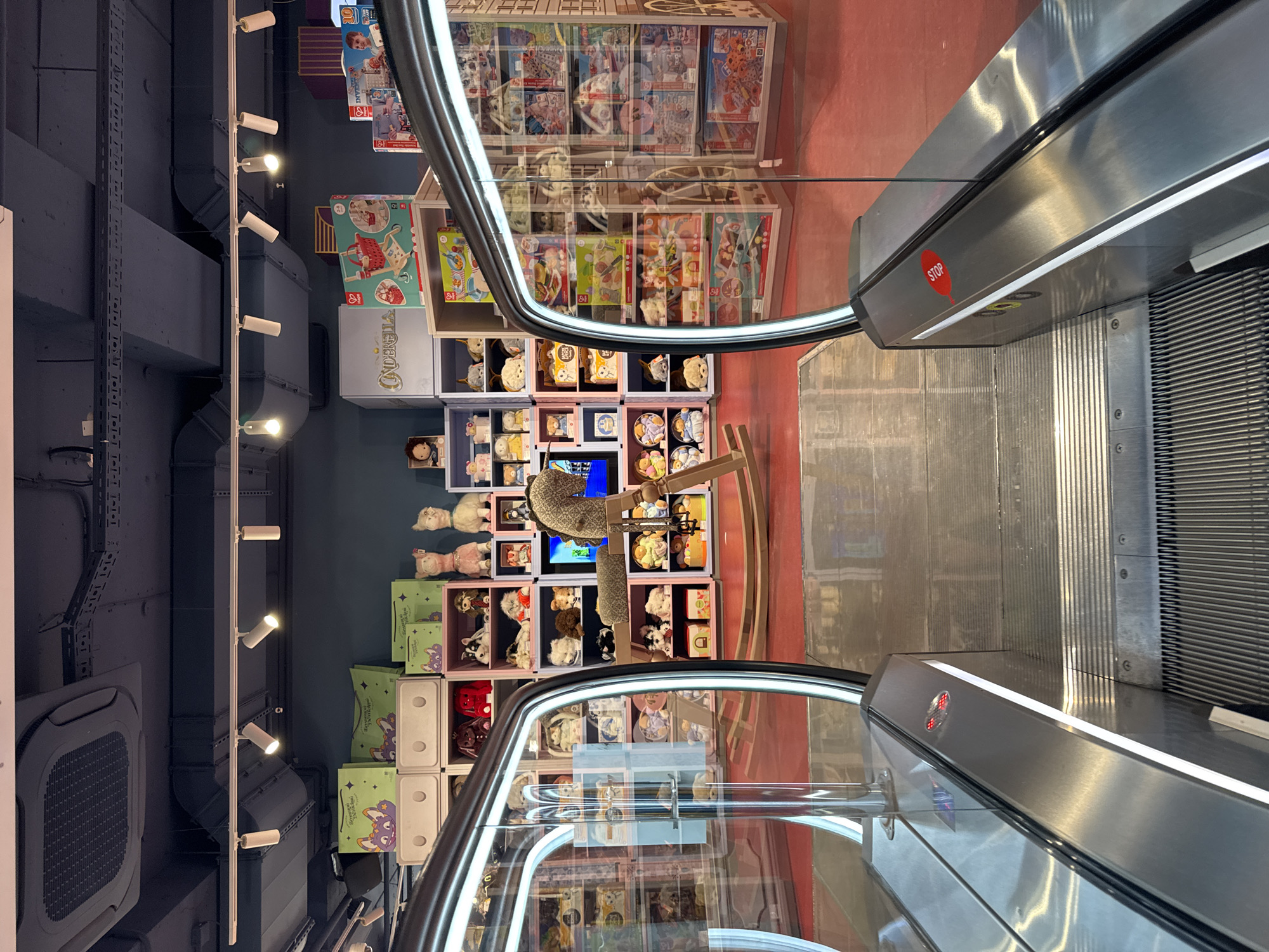 The image shows an indoor toy store, likely a modern department store, with escalators leading upwards. Shelves are filled with stuffed animals and boxed toys. There is a wooden rocking horse in the center of the image. The scene is brightly lit, with track lighting visible on the ceiling. The red floor indicates a commercial space. The image does not provide any specific information about the city of Yekaterinburg, Russia.