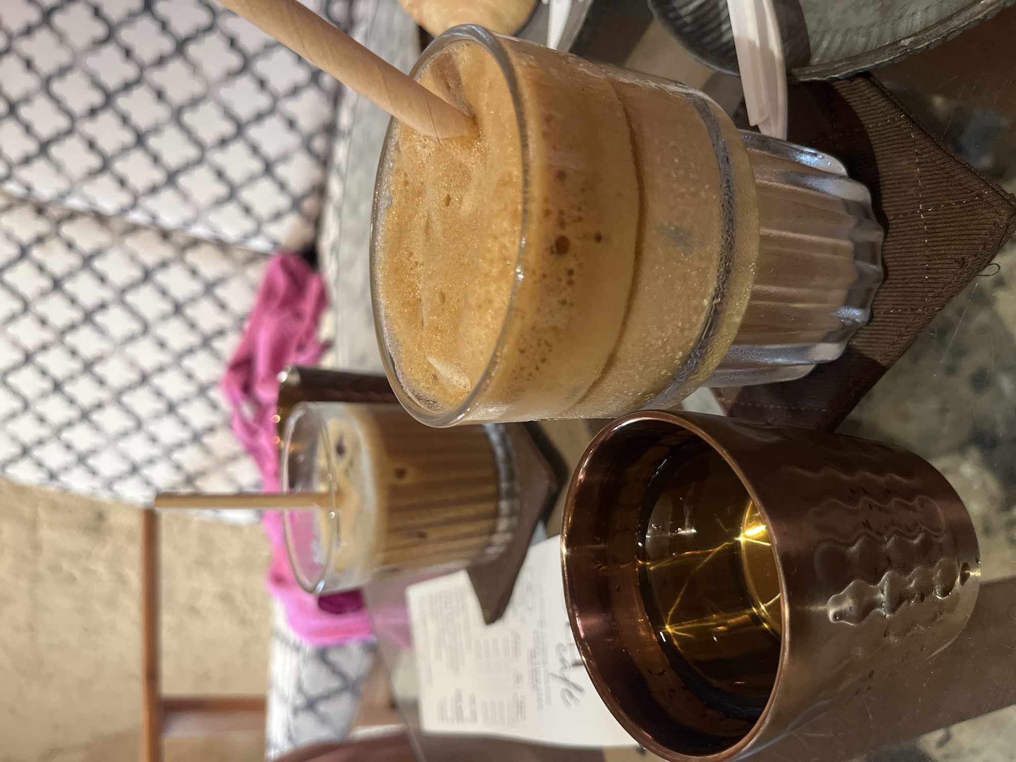 The image captures a close-up of a table setting in what appears to be a cafe. Dominating the scene are two glasses filled with frothy, coffee-colored beverages. One glass has a straw inserted, and the drink exhibits a light brown, foamy texture. Adjacent to it is a copper-colored mug filled with a clear liquid, possibly water, reflecting light from the surroundings. Partially visible beneath the drinks is a menu or receipt from the cafe. The background features a blurred glimpse of a patterned cushion and a pink garment, indicating a seating area. The overall setting suggests a relaxed, casual atmosphere in a cafe in Unknown, Unknown.