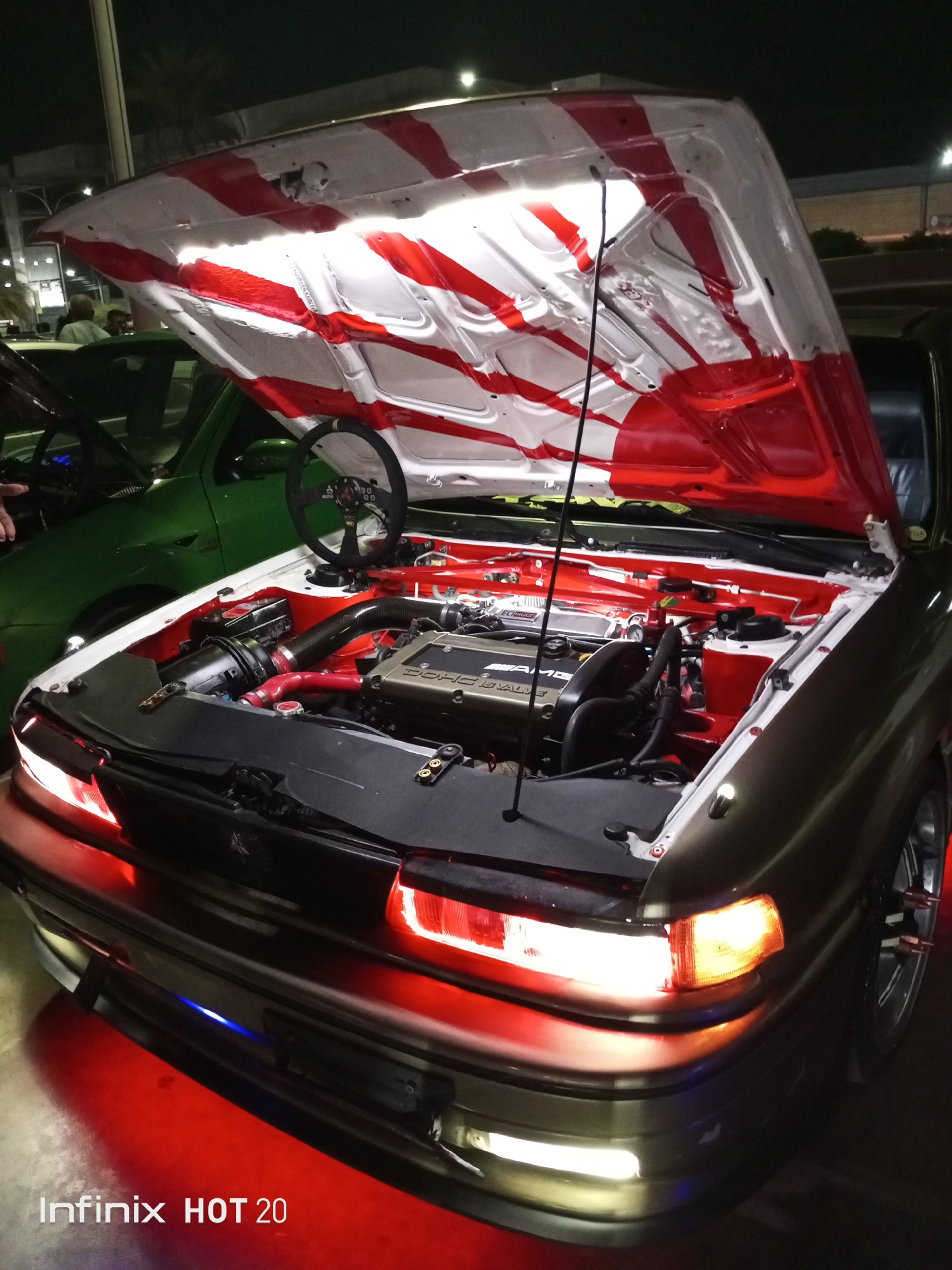 The image captures a modified dark-colored car with its hood open, revealing a customized engine compartment painted with a white and red "Rising Sun" motif on the underside of the hood. The engine itself is visible, along with other components in the engine bay, all painted red. A steering wheel is resting on the engine bay. The car features illuminated red lights around the front bumper and headlights. The photograph appears to be taken at night, possibly in an urban setting, indicated by building lights in the background and the presence of another car parked nearby. The text "Infinix HOT 20" is at the bottom of the picture, suggesting the model of the capturing device. The location details, Porlamar, Venezuela, are not directly visible in the image.