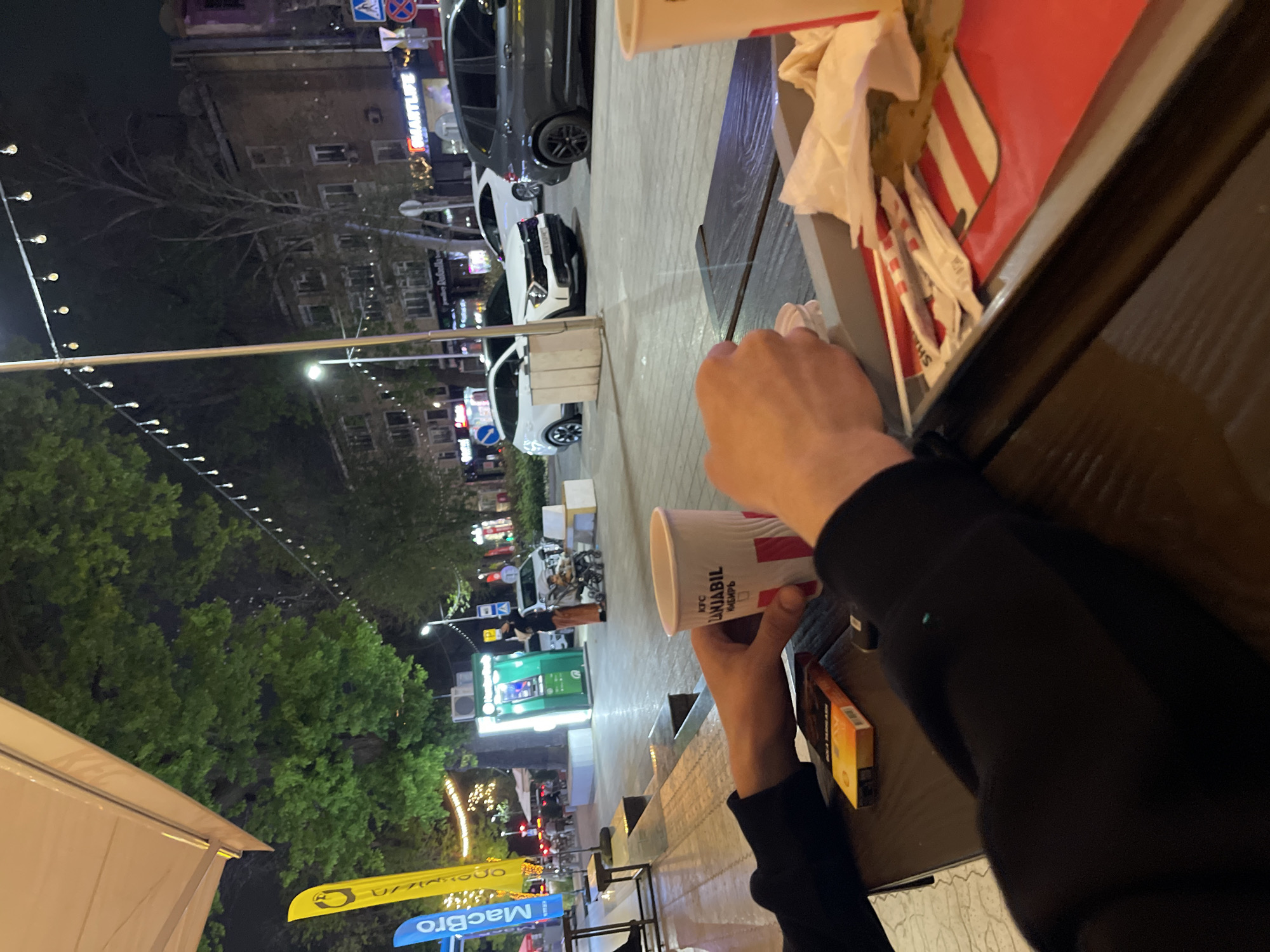 The image shows a person holding a KFC cup in an outdoor setting. The cup has the words "KFC," "JANIBIL," and "KIBIR" printed on it. The person is sitting at a table where a fast food order is placed, there are burger wrappers and other fast food items. In the background, there is an urban environment with cars, buildings, and street lights. A row of trees lines the street. The camera perspective is in a rotated vertical position, looking across the table and out toward the cityscape. Based on the text visible on a banner, the location may be Tashkent, Uzbekistan.