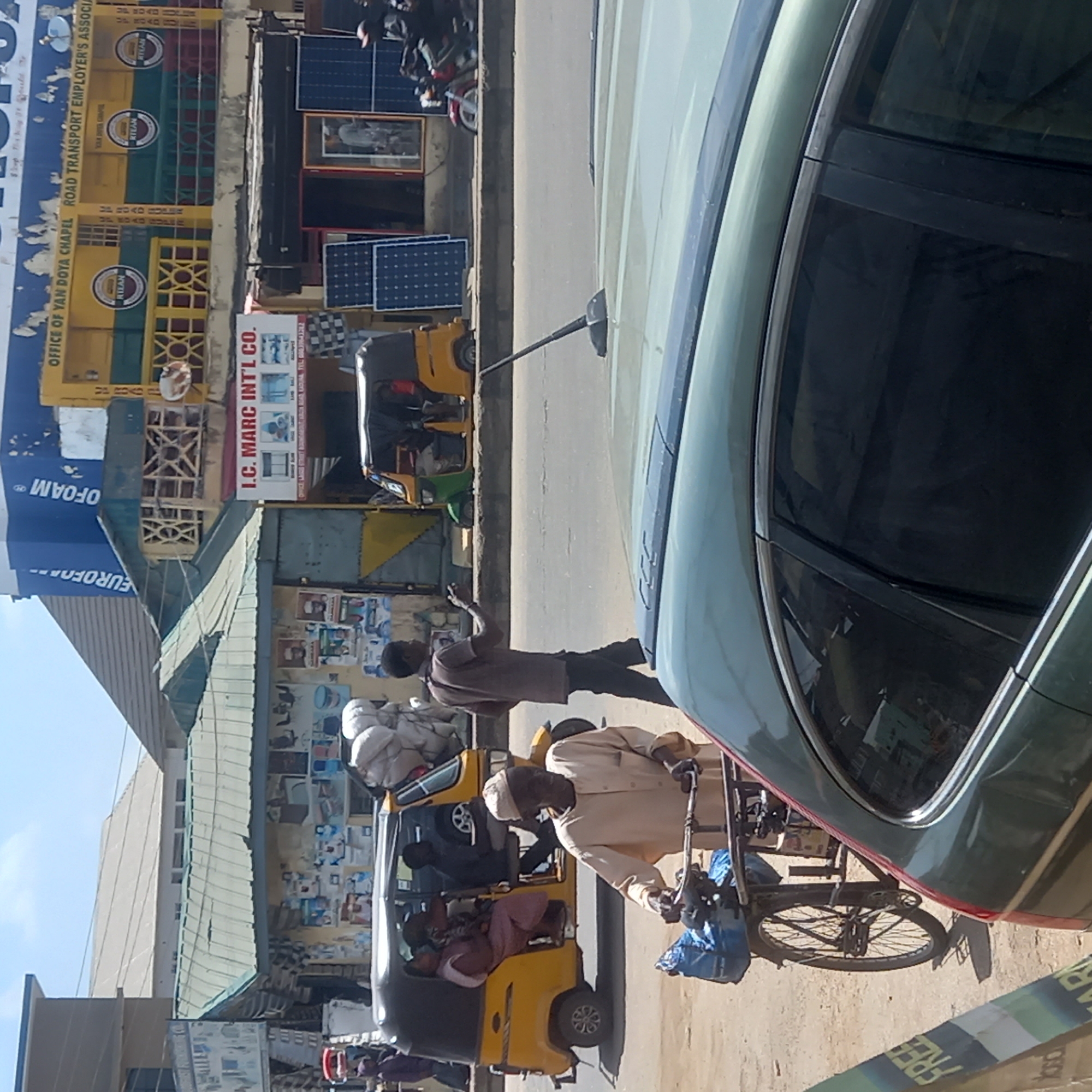 The scene captures a street view in what is likely the city of Dadi Riba, Nigeria, based on the text on a building in the upper left which reads "OFFICE OF YAN DOYA CHAPEL ROAD TRANSPORT EMPLOYER'S ASSOCIA" and "I.C. MARC INT'L CO.". The image is taken from the perspective of someone inside a car, with a partial view of the car's side. The street has various vehicles, including two auto rickshaws (tuk-tuks), one of which is carrying passengers and supplies. A man walks next to the rickshaw, pointing towards the road. Another individual is riding a bicycle or tricycle with a carrier attached, possibly with packages or bags.
The buildings lining the street have storefronts, some displaying signs or posters. Solar panels are visible above one of the buildings. One building has signage displaying "I.C. MARC INT'L CO.", while another building has an yellow and green facade. The overall scene suggests a bustling urban environment.