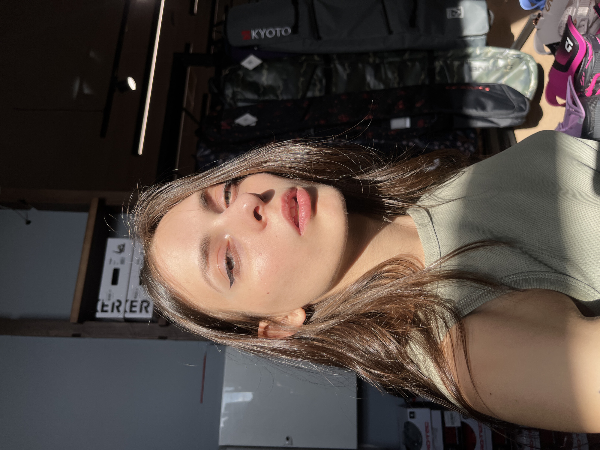An indoor scene within a retail establishment, specifically a sports equipment or outdoor gear store, likely a branch of Trial-Sport (Триал-Спорт) in Krasnoyarsk, Russia. The primary subject is a young adult female, positioned centrally and looking directly at the viewer. She has long, dark brown hair and is wearing a light green ribbed tank top. Her face features visible winged eyeliner and slightly parted lips. Strong, directional sunlight illuminates the right side of her face and torso, casting a deep shadow across her left side.
In the left background, a light-colored wall or display panel is partially visible, reflecting light. Below it, several white product boxes are stacked, one showing the partial text "ERIER" on its side. The right background displays multiple dark-colored bags or cases, consistent with sports equipment covers or travel bags, hanging from racks. One bag features "KYOTO" written vertically in white on a red patch. Other bags exhibit camouflage patterns. The ceiling above is dark, fitted with modern track lighting fixtures. In the lower right foreground, a display surface holds various pieces of brightly colored sports equipment, predominantly in pink and purple, which appear to be protective gear or specialized footwear, with at least one item bearing a "C1" logo.