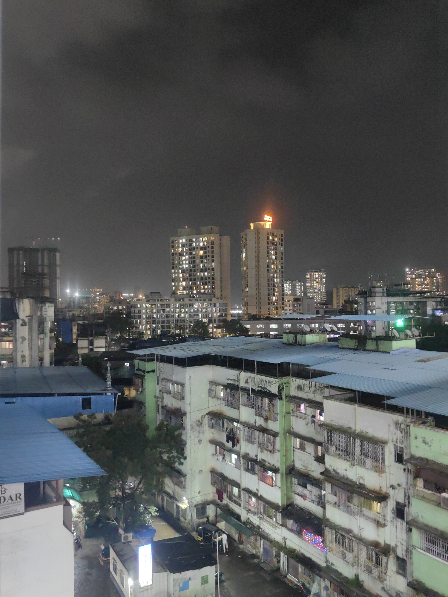 This is a nighttime, high-angle photograph of an urban environment in Mira-Bhayander, India. The scene is dominated by a dense collection of residential buildings of varying heights. In the foreground, a multi-story apartment building with a pale green facade is visible, featuring numerous balconies with metal railings and windows. Laundry is hung from some of the balconies. Adjacent to this building, to the left and slightly further back, is another apartment building with a white facade, also displaying multiple windows and balconies. To the left of these buildings, a section of a blue-roofed structure is visible, and a sign with the text "B DAR DEVELOPERS LLP" can be seen on its side.
In the midground, several other apartment buildings are illuminated by interior lights, creating a pattern of lit windows against the dark sky. Two taller, modern buildings stand out in the distance; one is a light-colored skyscraper with many illuminated windows, and a bright orange illuminated sign is visible at its summit. Another similarly tall building with lit windows is located to its left. Further in the distance, more buildings with illuminated windows extend into the hazy night.
The sky is dark and overcast, with some subtle variations in shade indicating cloud cover. The overall impression is of a densely populated urban area at night, with the lights of residences and commercial establishments illuminating the scene. The ground level is partially visible in some areas, showing what appears to be a street or alleyway with a few parked vehicles, including what looks like an auto-rickshaw. A few trees are also visible, their foliage dark against the lit buildings. The image captures the bustling atmosphere of city life after dark.