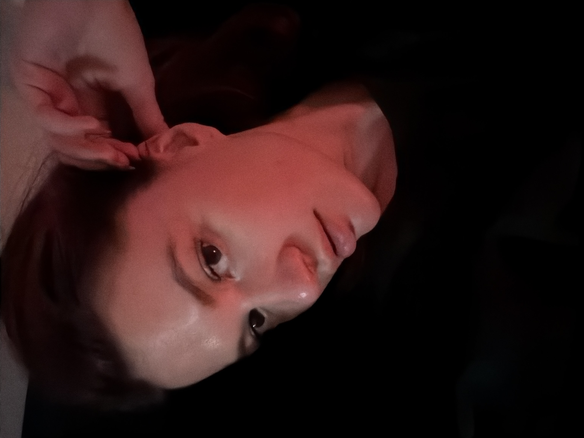 A close-up, tilted portrait shows a fair-skinned person, possibly female, in near darkness. The person's face, lit with a reddish hue, is turned towards the lower left. One of the person's hands, also lit with the same red hue, is near their ear. The background is predominantly black, obscuring any specific environmental details. There are no details available to identify the location as Belgrade, Serbia.