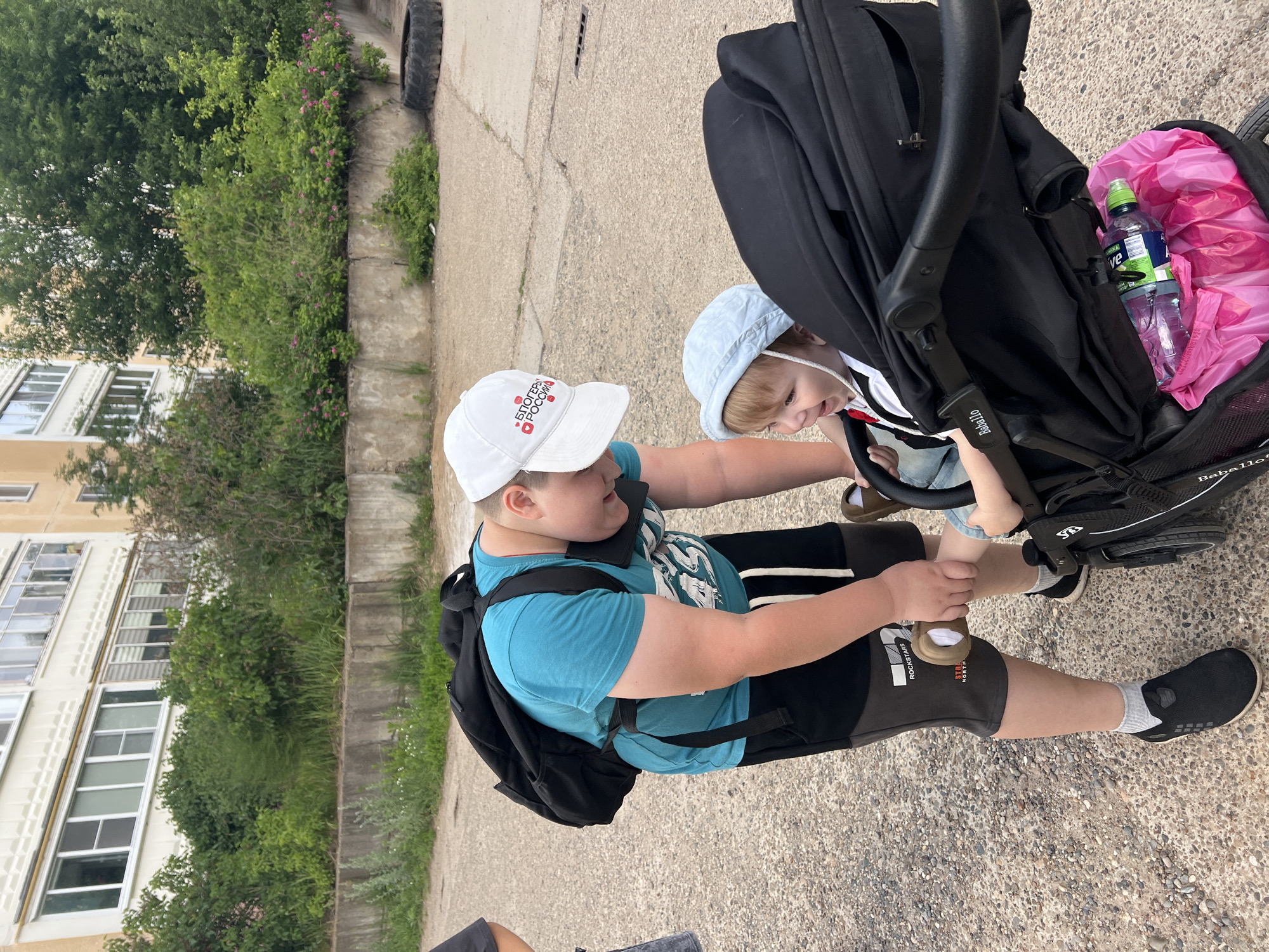 In Noviy, Russia, a young boy wearing a blue t-shirt, black shorts, and a white baseball cap with Russian text is leaning over a black stroller. He is holding the foot of a baby who is seated in the stroller. The baby is wearing a light blue sun hat and appears to be smiling. The background features a concrete wall with green bushes and trees, and a multi-story building with windows. The ground is paved with gravel and concrete.