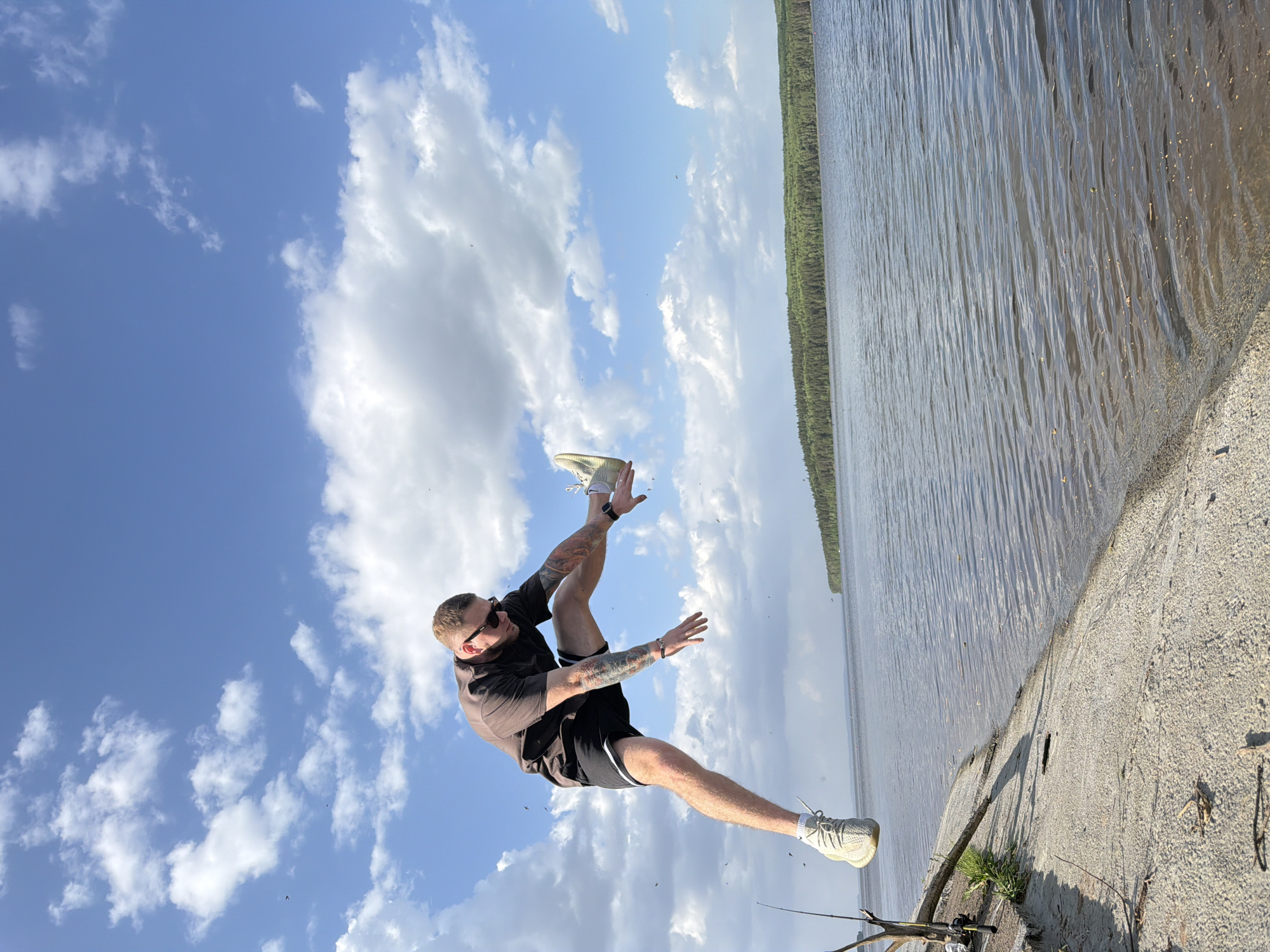 A man is shown outdoors appearing to be falling or stumbling. He wears a dark shirt, black shorts with white trim, sunglasses, and light-colored sneakers. He has tattoos on his arms and fair skin. He is airborne, with one leg extended and the other bent at the knee. His arms are outstretched for balance. The scene includes a body of water, possibly a river or lake, with a forest visible on the opposite shore. A concrete edge suggests a pier or embankment. To the lower right is a fishing rod. The background sky is blue with scattered clouds. Based on the presence of a pier-like structure along a body of water with forest visible on the opposite shore, this might be a body of water on the Kama river, likely near the city of Chaikovsky, Russia.