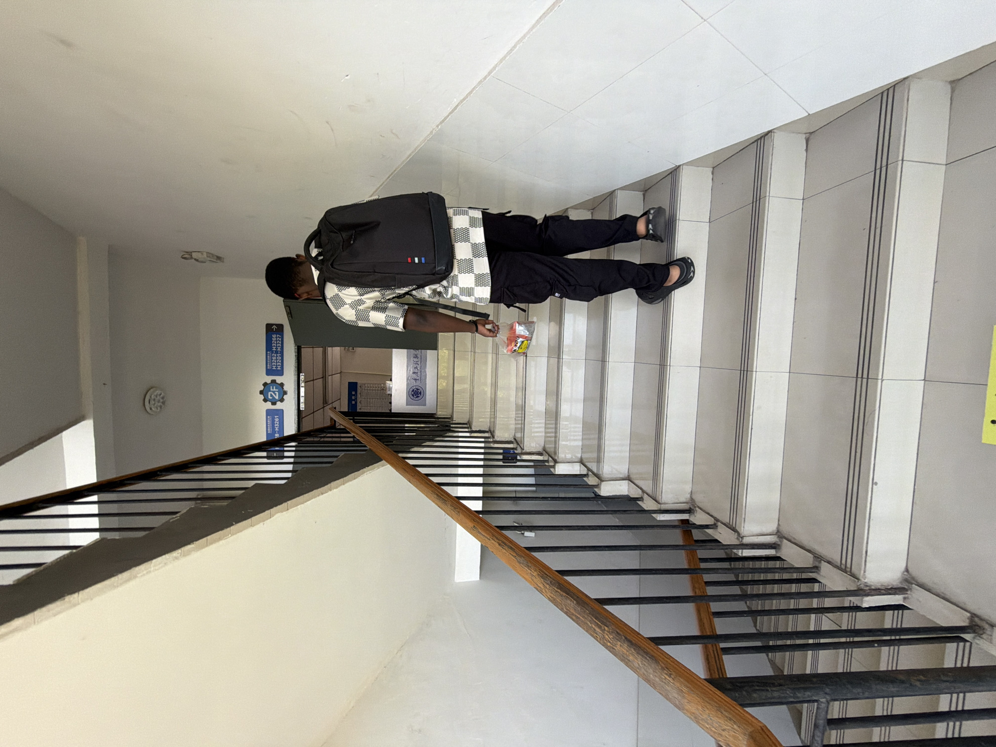 A Black male wearing a checkered shirt, black cargo pants, and black slip-on shoes walks up a white tiled staircase. He has a black backpack on and is holding a clear plastic bag containing items in his left hand. The staircase has a wooden handrail on the left and black metal railings. Signs are visible at the top of the staircase, including one with "2F" and Chinese characters. The walls are painted white, and there is a fire alarm on the ceiling. The environment appears to be an indoor staircase in a building, possibly a university or office building, located in Jiangjin District, Chongqing, China.