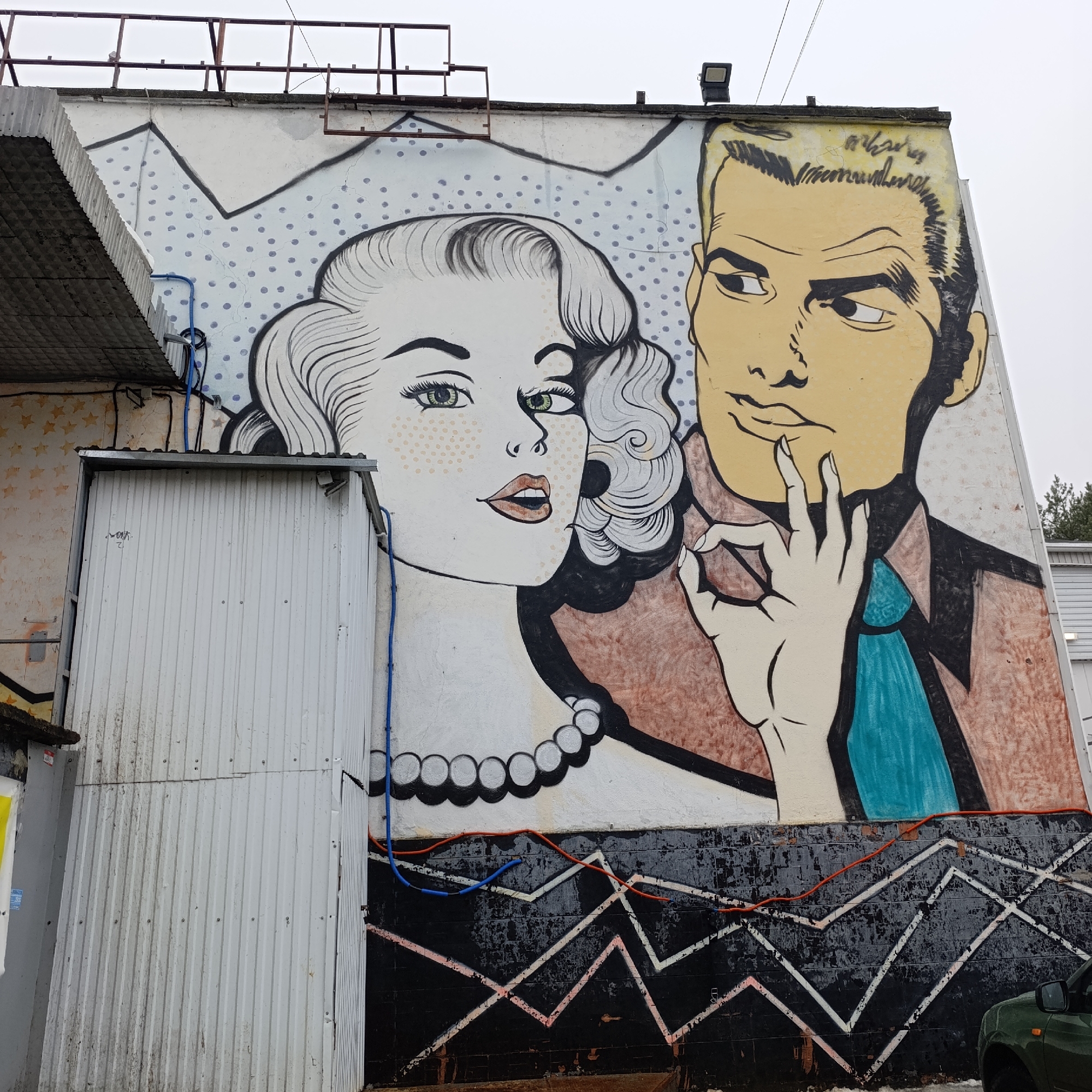 The image shows a mural on the side of a building in Chaikovsky, Russia. The mural depicts a stylized couple in the style of 1950s or 1960s comic book art. The woman has wavy blonde hair, green eyes, and a pearl necklace. The man has blonde hair, a brown suit jacket, and a teal tie. He is making an "okay" gesture with his hand near his face. The background of the mural is a light color with darker gray polka dots and zig-zag patterns at the bottom. The mural is on a wall, which also has visible pipes. A corrugated metal shed or structure is attached to the side of the building. A vehicle can be seen on the right edge of the image.
