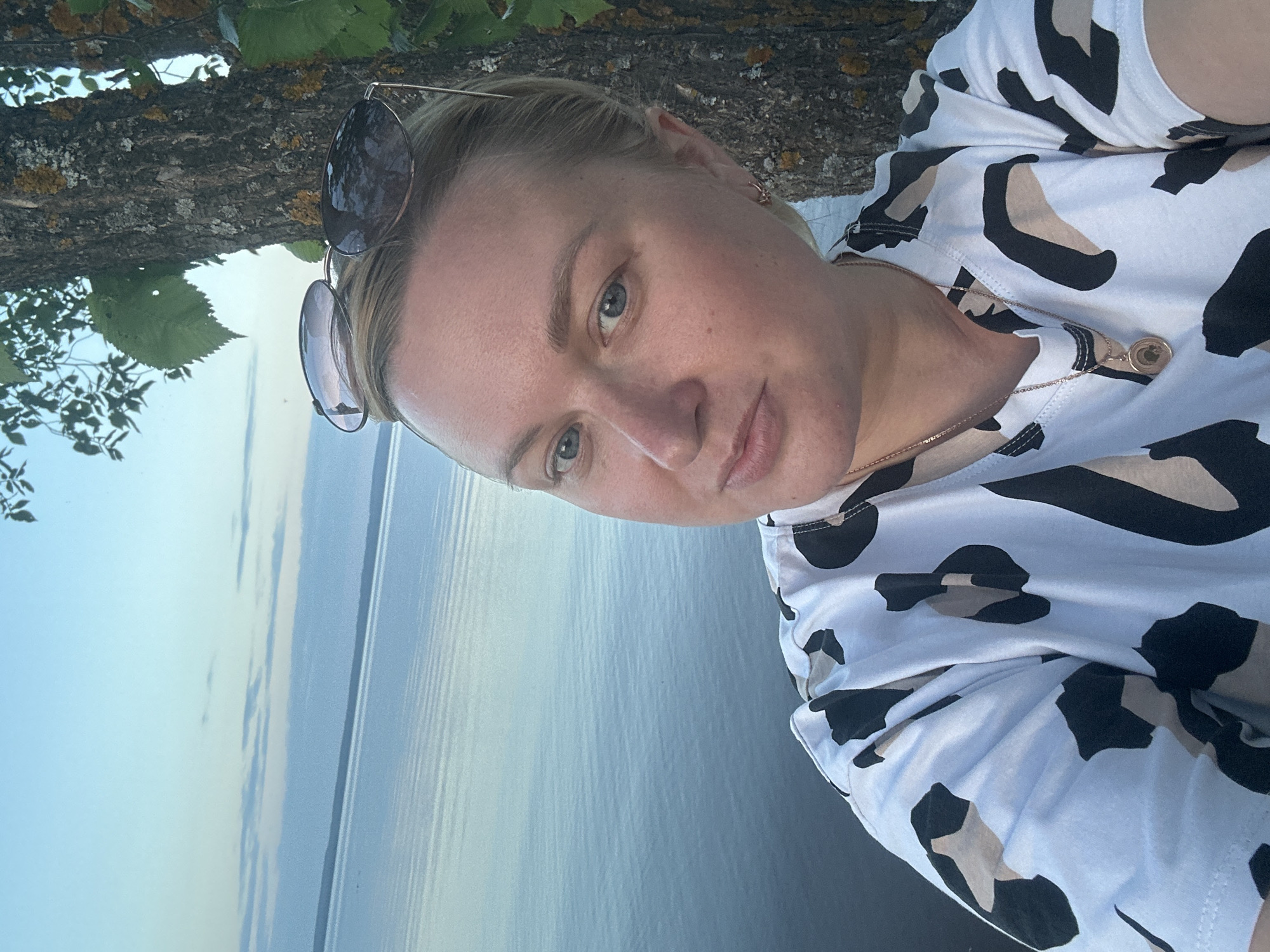 A woman is taking a selfie by a large body of water. She has blonde hair pulled back and sunglasses on her head. She is wearing a white t-shirt with a black and beige leopard print. She is also wearing a delicate gold necklace with a round pendant. Behind her, a tree with mossy bark and green leaves is visible, as is a calm body of water with a pale blue sky overhead. The scene suggests a serene outdoor setting, possibly near a lake or river. There is no indication of Noviy, Russia, in the image.