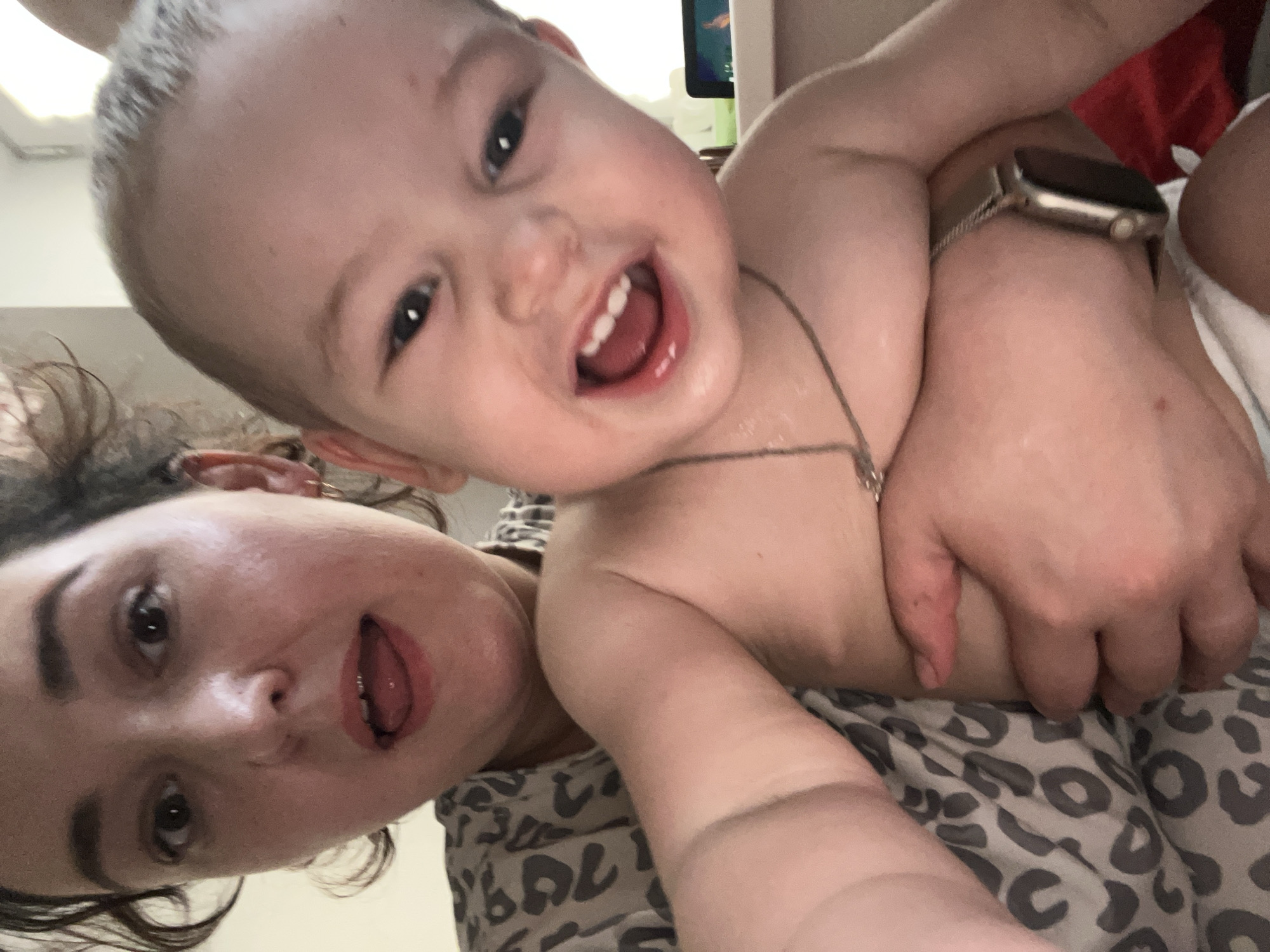 This is a close-up selfie of a woman holding a baby. The woman is on the left side of the frame, her face is partially visible, and she is sticking her tongue out. The baby is in the center and right side of the frame, looking at the camera and smiling widely with its mouth open. The baby is shirtless and wearing a silver necklace and a smartwatch on its left wrist. The woman is wearing a gray and black leopard-print shirt. The background is blurred but appears to be indoors, possibly a room with a window. There is no identifiable information in the image that would indicate the location in Chelyabinsk, Russia.