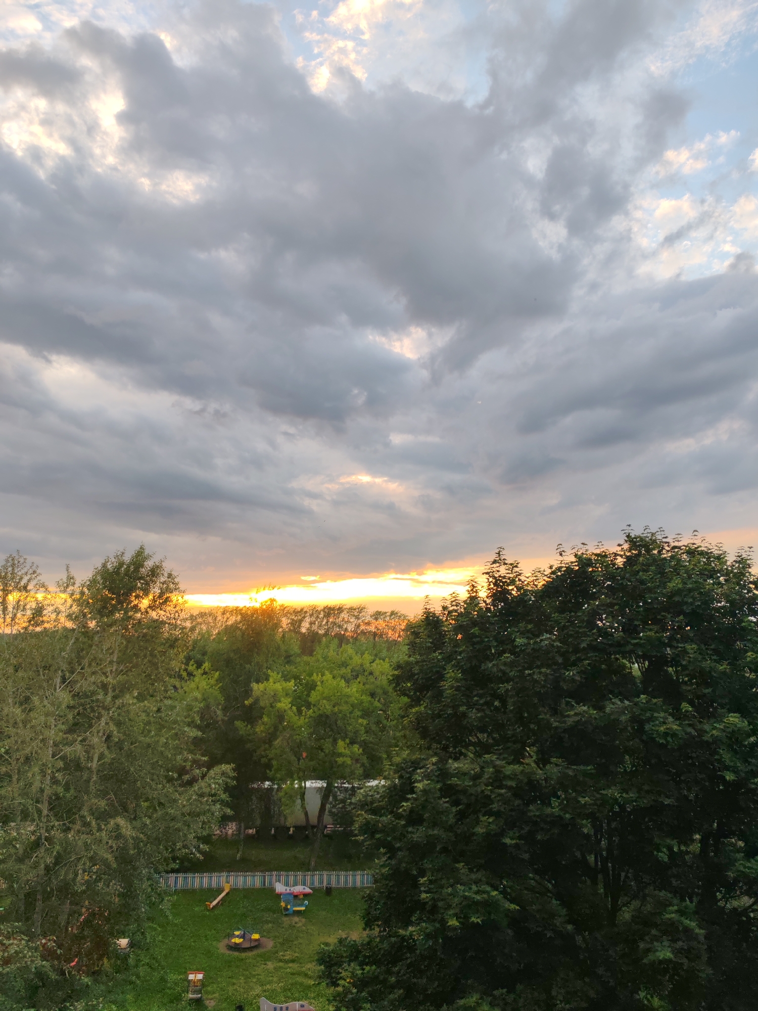 The image captures a dusky sky with a band of golden light from a setting sun illuminating the horizon, peeking through a heavy layer of grey clouds. Below the sky, a dense line of green trees forms a horizon, with some trees closer and larger, appearing dark green and full of foliage. In the foreground, a grassy area is visible with children's playground equipment, including a see-saw, a spinning ride, and a small airplane-shaped play structure. A colorful striped fence encloses part of the playground. A trailer is visible behind the fence, partially obscured by trees. The scene suggests a residential area or park within the city of Сокол, Russia, likely during late afternoon or early evening.