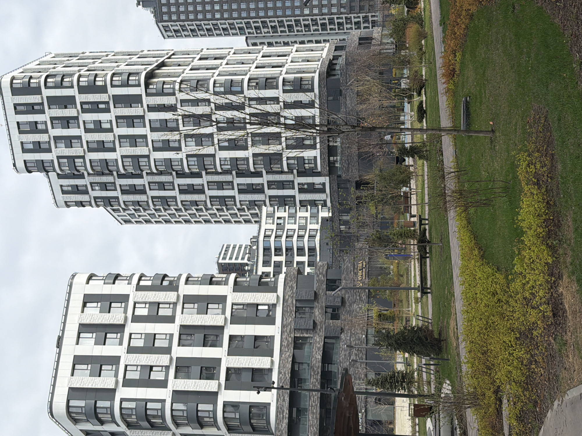 The image shows a modern urban landscape with several high-rise buildings in Перовское сельское поселение, Ukraine. The buildings are characterized by a distinctive black and white patterned facade with rounded edges. A park area is visible in the foreground, featuring a green lawn, trees, shrubs, and a pedestrian walkway. Some benches are also present in the park. The sky is overcast.