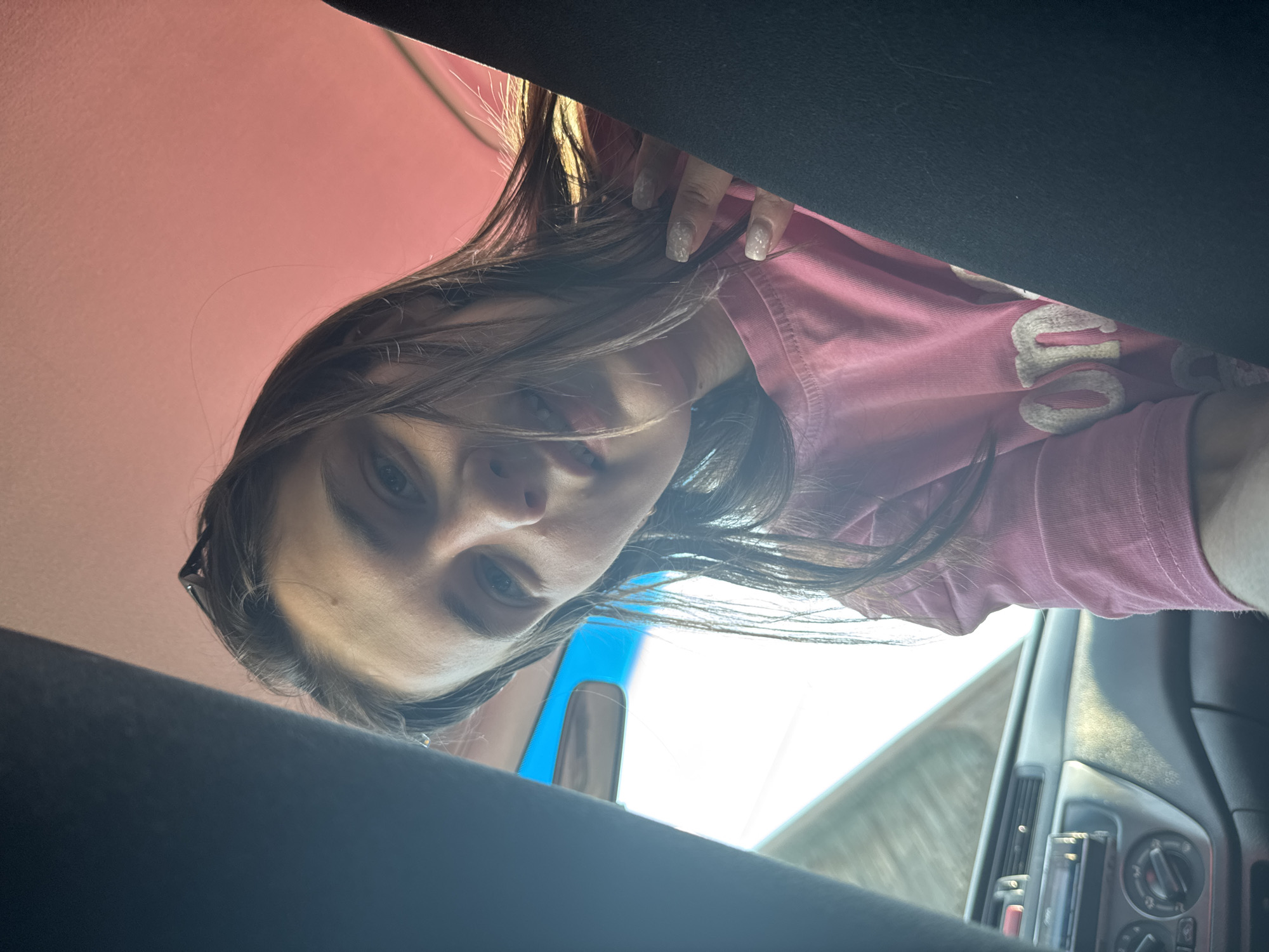 A woman with long brown hair and pale skin, wearing a pink shirt, leans over the camera inside a vehicle. Her nails are manicured with a glittery finish. The interior of the vehicle is visible, including the dashboard, window, and roof. The location is unknown; there is no evidence to suggest the scene is in Chaikovsky, Russia.