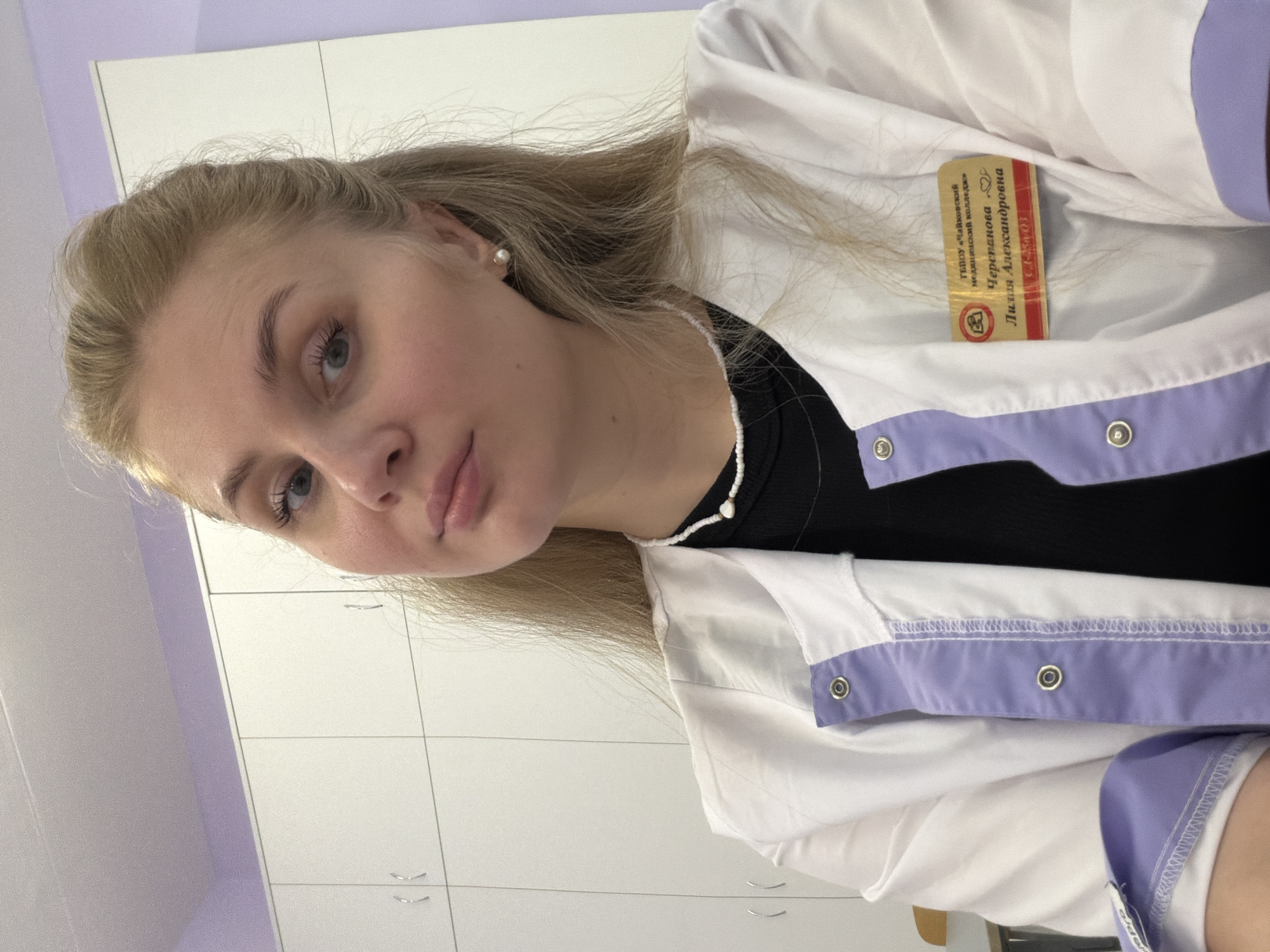 A young woman with blonde hair, styled partly up, is depicted from the chest up, looking directly at the camera. She wears a white lab coat with lavender accents on the lapels and cuffs, over a black top. A white beaded necklace and a single pearl earring on her right ear are visible. Attached to the right lapel of her lab coat is a gold-colored nametag with red trim. The nametag reads in Cyrillic: "ГБПОУ «Чайковский медицинский колледж»" (State Budgetary Professional Educational Institution "Chaikovsky Medical College"), followed by "Черепанова Лилия Александровна" (Cherepanova Liliya Aleksandrovna) and a code "ГС-14-03". The background consists of white cabinets with light-colored handles and a section of a light purple wall above them. The scene is located within the Chaikovsky Medical College in Chaikovsky, Russia.