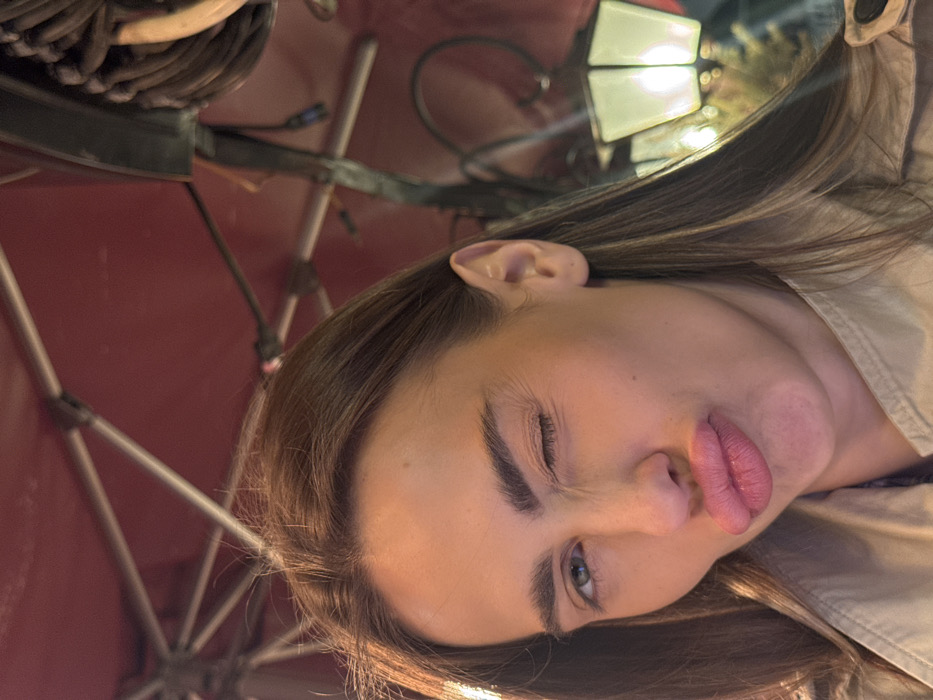 The image presents a close-up, frontal portrait of a female individual in an outdoor or semi-outdoor setting. The subject, with medium-length brown hair, is wearing a light-colored, possibly tan, trench coat. Her left eye is closed in a wink, and her right eye is open. Her lips are pursed into a puckered expression. The background features a reddish overhead canopy or awning structure supported by light-colored metal framing. To the right of the subject, an illuminated white, multi-paned lantern-style light fixture is visible, emitting a warm glow. Additional indistinct dark bundled objects or wiring are present in the upper right background. The scene appears to be set at night or in low light conditions, consistent with an outdoor seating area or cafe in the city of Venice, Italy.