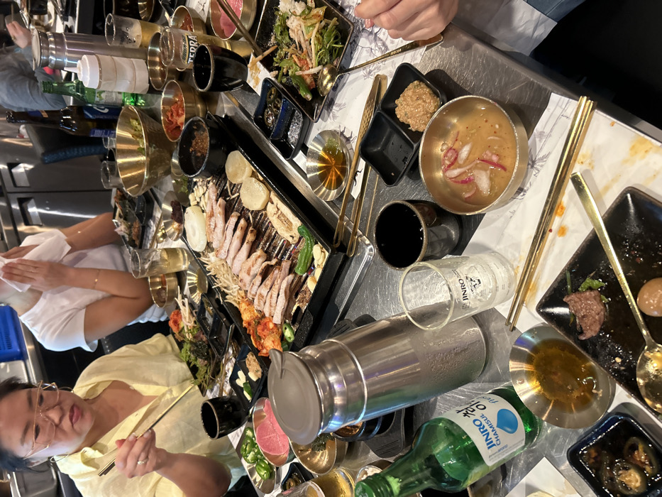 A Korean barbecue meal in progress. A grill with pork belly and vegetables dominates the table, surrounded by small dishes of kimchi, sauces, and other banchan. Jinro soju bottles, a steel pitcher, and shot glasses suggest a convivial atmosphere. Two women and a person wearing a white shirt are enjoying the meal, one actively using chopsticks. Overall, the table is filled with food and drinks.