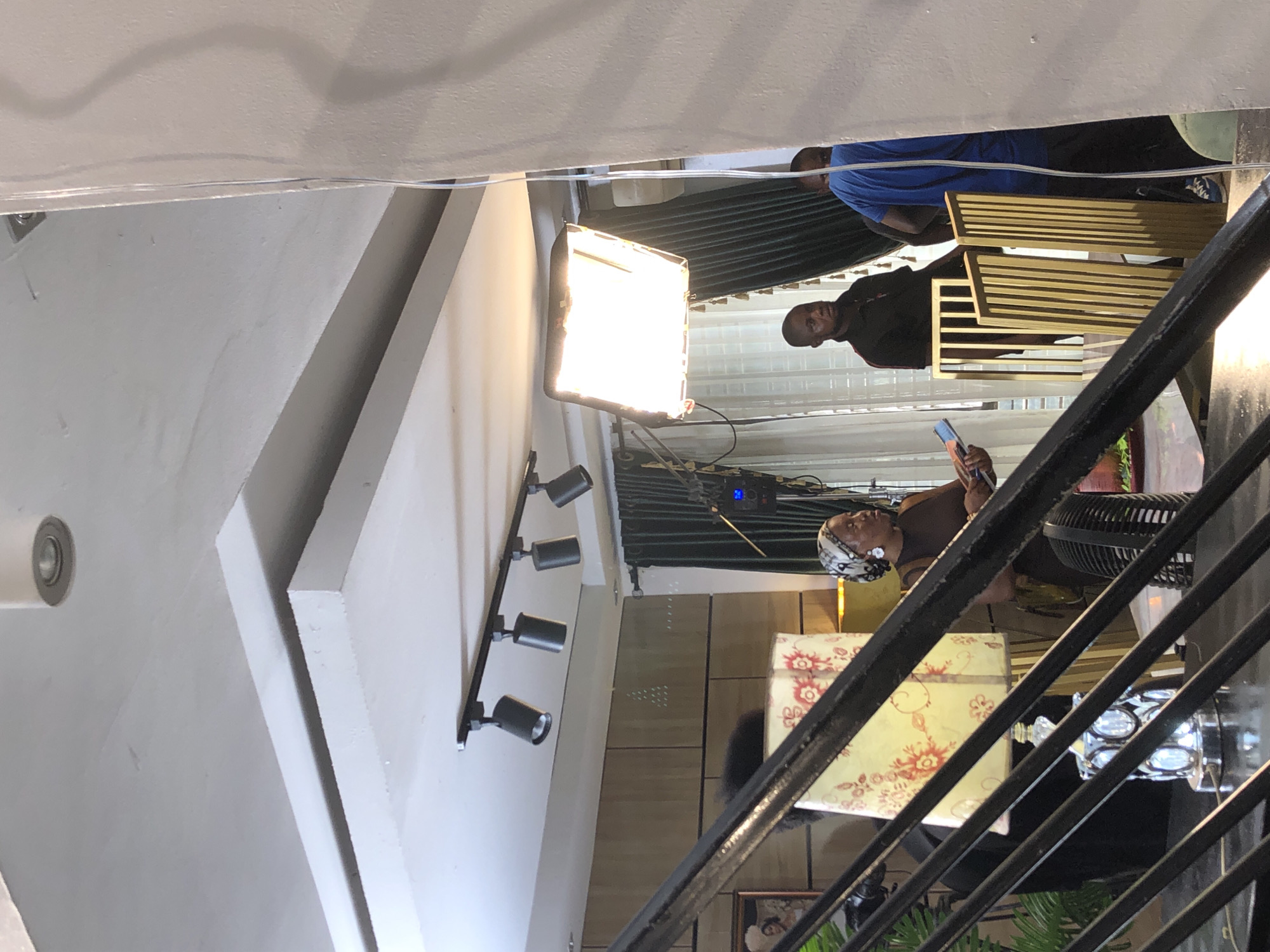 The image captures an indoor scene, likely a film or photo shoot, viewed from an elevated perspective looking down through a black metal railing. The central focus is a large, bright rectangular artificial light source (softbox) illuminating the area.
Three individuals are visible. On the right side of the frame, partially obscured by the railing, an older woman with a patterned white headscarf and a dark top is holding a smartphone, possibly taking a picture or video. Behind her, a black fan is visible, and next to her is a decorative yellow box with red floral patterns.
Further back and to the left of the woman, two men are standing near a set of golden slatted chairs or benches. One man, wearing a black top, is more clearly visible, facing towards the right side of the frame. Another man, wearing a blue t-shirt and dark pants, is partially seen behind the golden furniture.
The setting features modern interior design elements, including a white ceiling with a recessed light and a wall-mounted track light fixture with four black spotlights. The background appears to have vertical blinds or slatted panels. A device with a small blue light can be seen near the top edge of the bright light source, possibly a controller or power pack. The overall lighting suggests an intentional, staged setup, characteristic of a professional media production. No discernible text is visible.