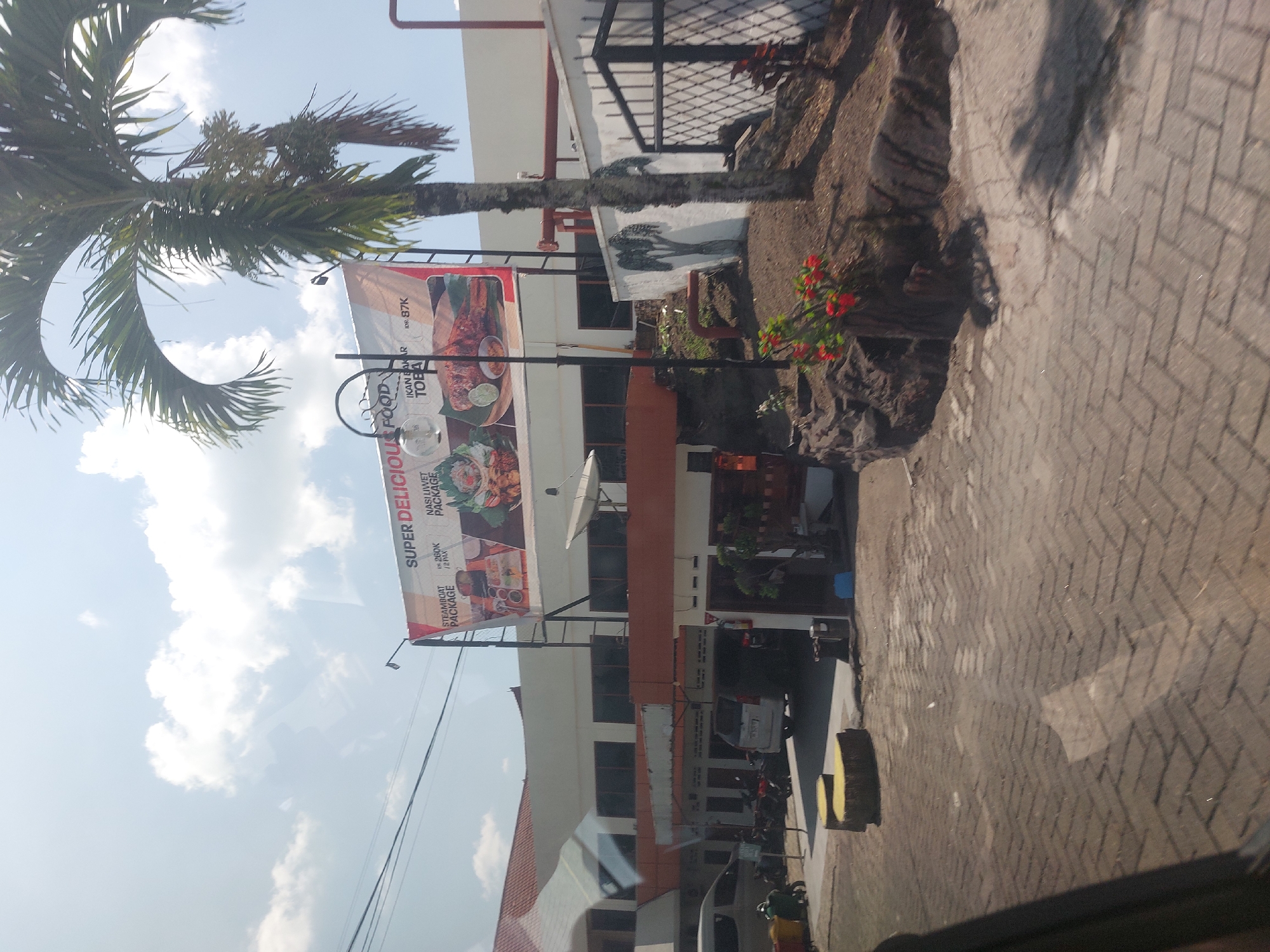 The image depicts the exterior of a restaurant building located in Tiga Raja, Indonesia. The building appears to be two stories high with white walls and orange accents. A large banner advertising "SUPER DELICIOUS FOOD" is prominently displayed on the front of the building, featuring images of various food packages and prices. A palm tree stands to the right of the banner, partially obscuring it. In front of the building, there is a paved area with parked vehicles, including a white van and several motorcycles. There are also some decorative elements, such as stone-like structures and a small patch of flowers. The sky is partly cloudy, indicating daytime.