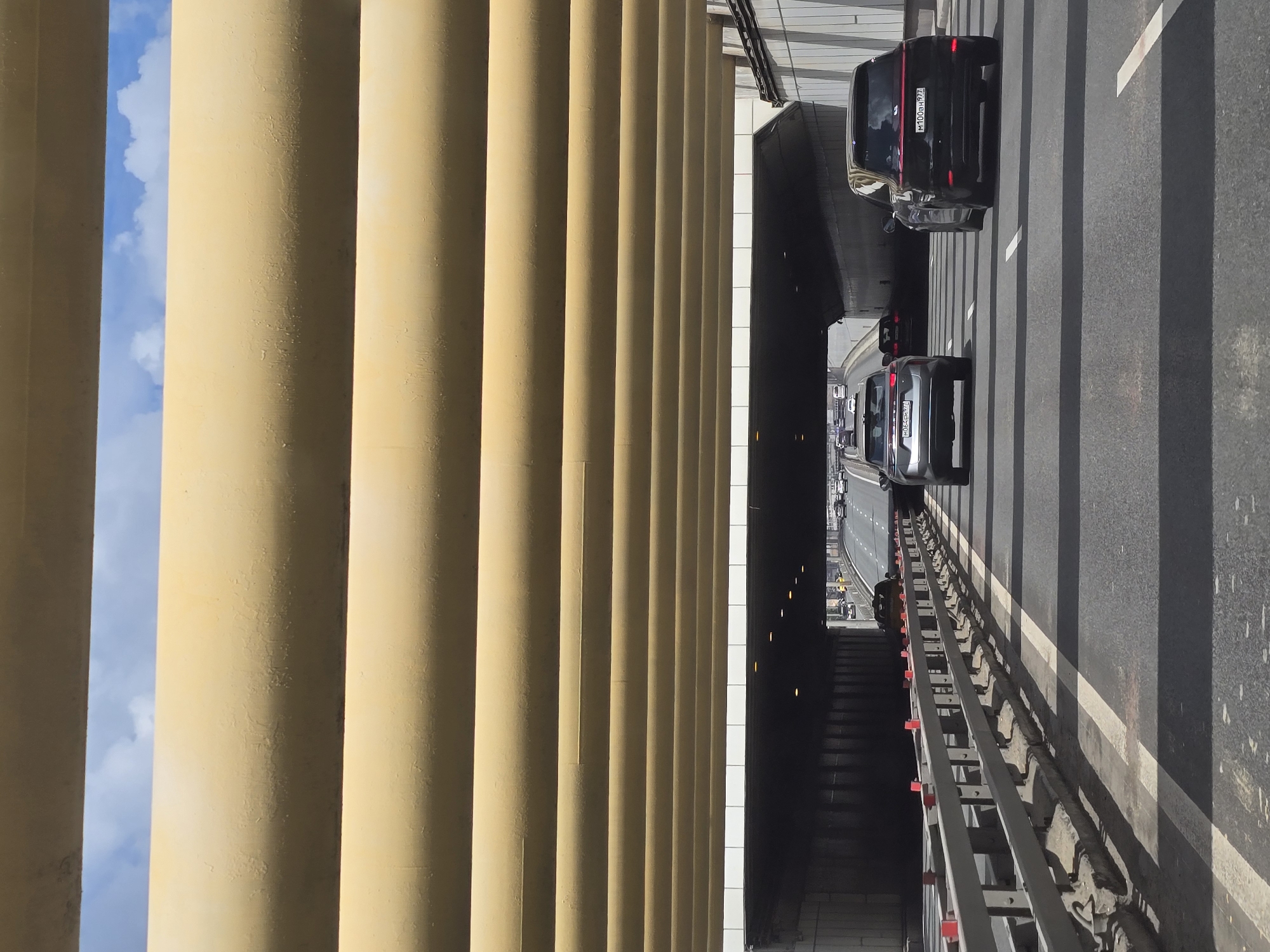 The image shows a segment of the Third Ring Road in Moscow, Russia, as viewed from a pedestrian overpass. On the left, a row of large, yellow cylindrical columns provides a screen. Traffic is visible on the multi-lane highway, with several cars in view. One car is black and another is silver. A pedestrian stairway descends from the overpass toward the road below. The scene is bathed in daylight, with a glimpse of blue sky visible between the columns on the left.