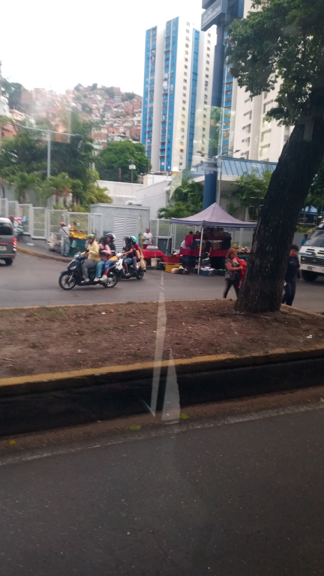 The image depicts a street scene in an urban environment, likely taken from inside a vehicle. Two motorcycles, each carrying multiple passengers, are visible on the road. Adjacent to the road, there is a makeshift market under a tent, with several individuals standing around it. In the background, tall buildings with blue and white facades are visible, along with a hillside covered in dense housing. A large tree is situated on the right side of the frame, partially obscuring the scene. A curb separates the road from a patch of dirt and vegetation. The lower portion of the frame shows the interior of a vehicle, including a portion of the window frame and the street surface. The location is Unknown, Unknown.