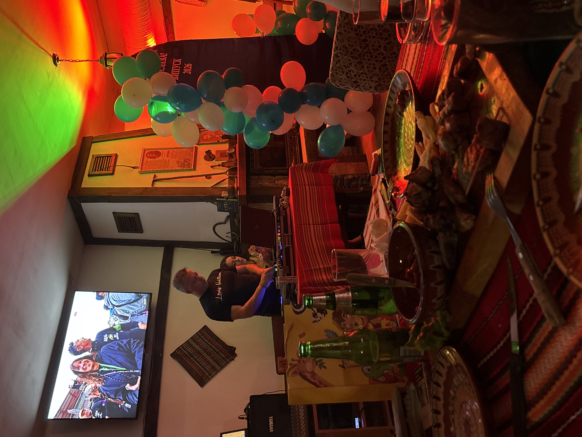 This image captures an indoor celebratory event occurring in a venue within Sofia, Bulgaria. The environment is characterized by dim illumination from prominent red and green spotlights cast onto the ceiling and upper walls.
In the mid-ground, a male DJ with short, grey hair is actively operating a sound mixer, with two laptops positioned on either side of the console. He is wearing a black t-shirt bearing the "Louis Vuitton" logo. Behind the DJ, a woman with dark hair is seated, focused on a personal electronic device. A black Yamaha speaker is visible to the left of the DJ setup. Mounted on the wall above the DJ is a large flat-screen television displaying an outdoor event, featuring a smiling woman in a dark blue jacket and a man in a black t-shirt among other individuals. A decorative woven fabric piece in a diamond shape hangs on the wall to the DJ's right.
On the right side of the room, a vertical banner in Cyrillic text displays "СЛАВА! ВИПУСК 2026" (Glory! Class of 2026). In front of this banner, a column of balloons, comprising white, teal, and orange spheres, serves as decoration. A brass lantern-style light fixture hangs from the ceiling in this area. A wooden cabinet or fireplace mantel below the banner features an antique-style framed portrait and other decorative items.
The foreground is dominated by a table draped with a striped red and orange tablecloth. Food items include a wooden serving board laden with grilled meat skewers (e.g., kebabs) and fried potatoes, and a brown ceramic bowl containing a red sauce. Beverages visible on the table consist of two green glass beer bottles, multiple clear drinking glasses, and dark ceramic mugs. Cutlery, including forks and knives, is laid out. A yellow gift bag adorned with cartoon animal illustrations rests near the beer bottles.