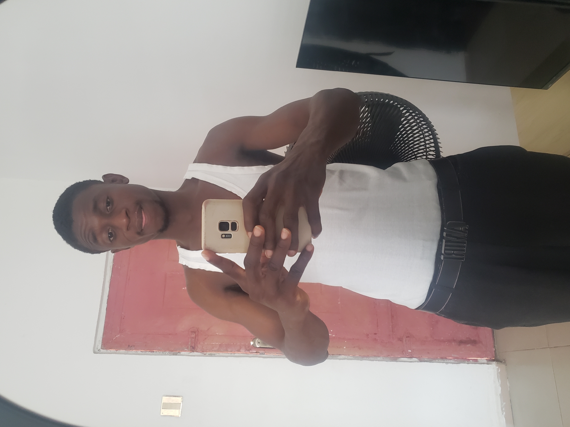 The image captures a young, dark-skinned man taking a mirror selfie indoors in Adonkolo, Nigeria. He is seen from the chest up, wearing a white tank top and black pants with a visible waistband. The man has short, dark hair and a slight, pleasant smile as he looks towards the smartphone he holds with both hands.
The background features plain white walls. To the man's left, a reddish-brown door, which appears somewhat worn, is visible. On his right side, a black object that resembles either a fan or the back of a chair is partially visible. Above this object, a large, black, rectangular screen, likely a television, is mounted high on the wall. Near the bottom left, a light-colored rectangular object, possibly an electrical outlet or light switch, is affixed to the wall. The floor in the foreground, visible in some crops, appears to be tiled.
The smartphone held by the man is light-colored and features a multi-lens camera setup on its rear. The waistband of his black pants has white text that, when viewed in the mirror, reads "AMUP," indicating the "PUMA" brand in reverse. The scene suggests a casual and personal moment captured within a simple indoor setting.