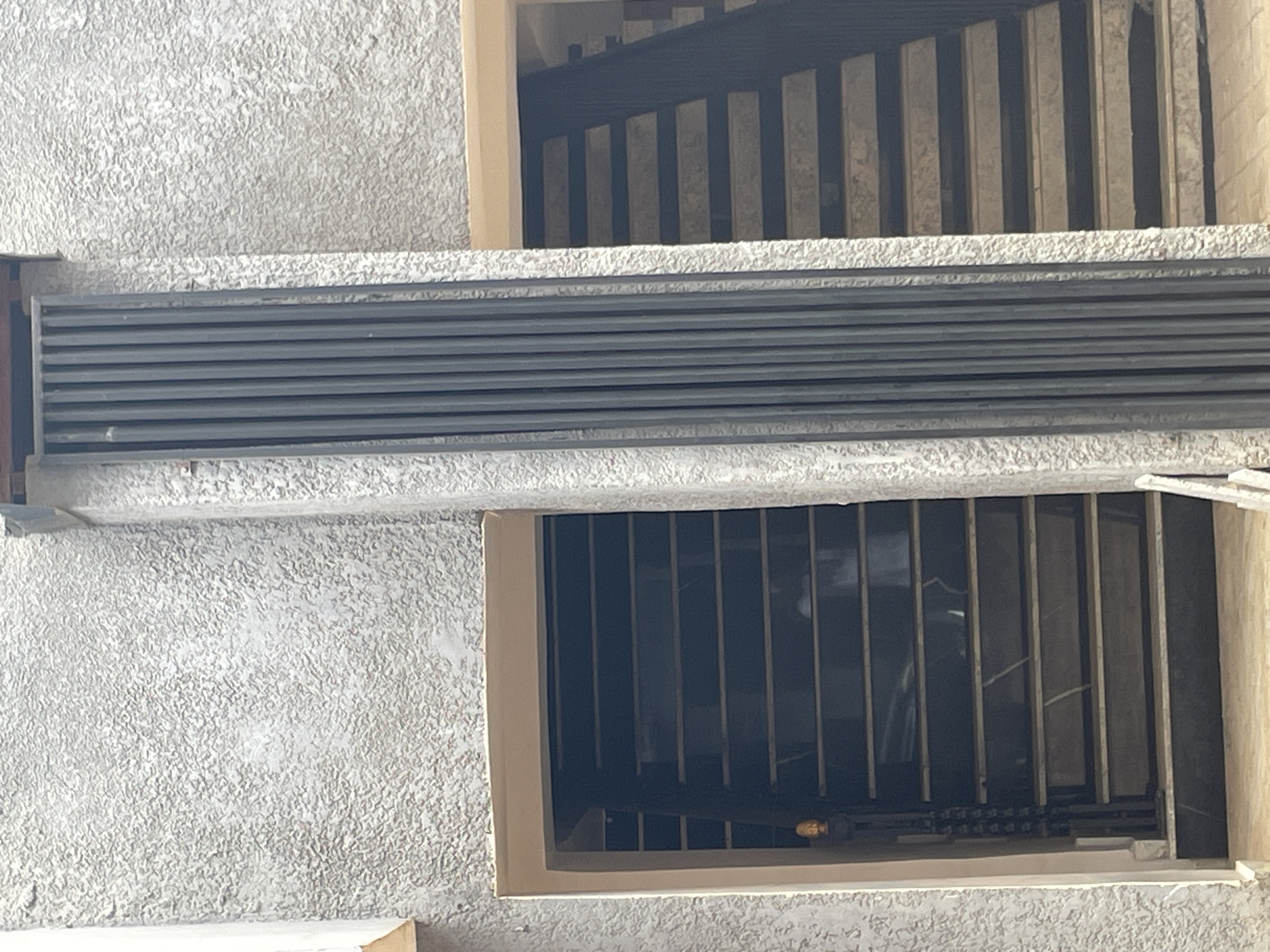 The image shows two outdoor staircases on a building with a textured, light gray facade. A vertical dark gray metal grate runs between the two staircases, extending from the top of the image downwards. The left staircase is dark, with what appears to be polished stone treads, and a wrought iron railing with a small golden light fixture at the bottom. The right staircase has lighter, possibly concrete treads, and a dark railing. The environment suggests an urban setting in Abuja, Nigeria, possibly an apartment complex or a public building. There are no people clearly visible, though faint shadowy figures can be discerned on the left staircase. The lighting is overcast, suggesting a cloudy day.