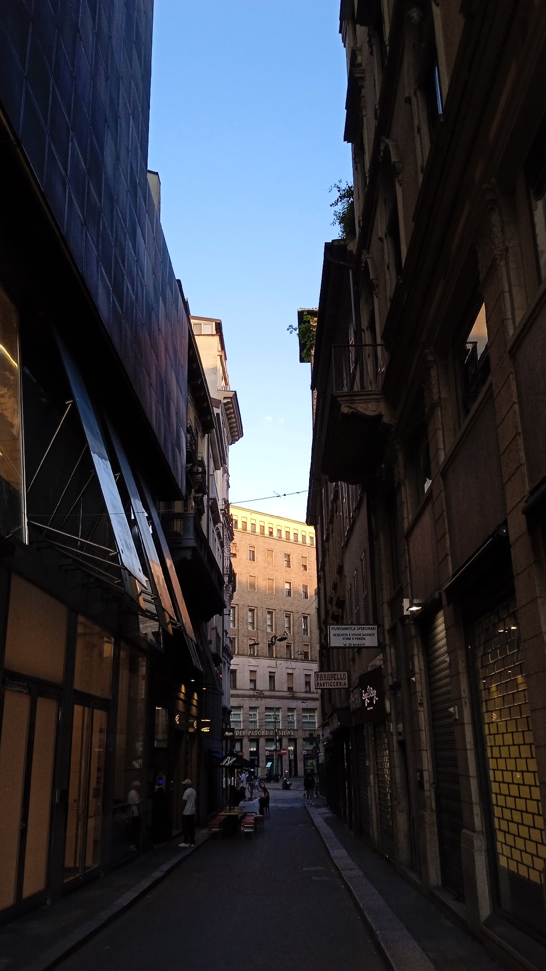 The image depicts a narrow street in Milan, Italy, with tall buildings on either side. The sky above is a clear blue, suggesting it's daytime. The street is paved, and there are shops and businesses lining its edges.
On the left side of the street, a modern building with large glass windows dominates the foreground. Its facade is dark and appears to be made of metal or a similar material. The windows reflect the sky and the opposite buildings. Further down the street on the left, there are more shop fronts, some with awnings. People are visible, standing and sitting at tables outside a cafe or restaurant.
On the right side, older buildings with more traditional architecture are present. These buildings have balconies and numerous windows. A sign for "NUMISMATICA SPERONARI" and "PASTICCERIA" is visible, indicating a numismatist and a pastry shop. Rolling metal shutters are partially covering some of the ground-floor businesses.
The street itself appears to be mostly empty of vehicles, with a few individuals walking. The perspective of the image creates a sense of depth, drawing the viewer's eye towards the end of the street where more buildings are visible in the distance. The lighting suggests the sun is casting shadows, indicating it's likely late afternoon or early morning.