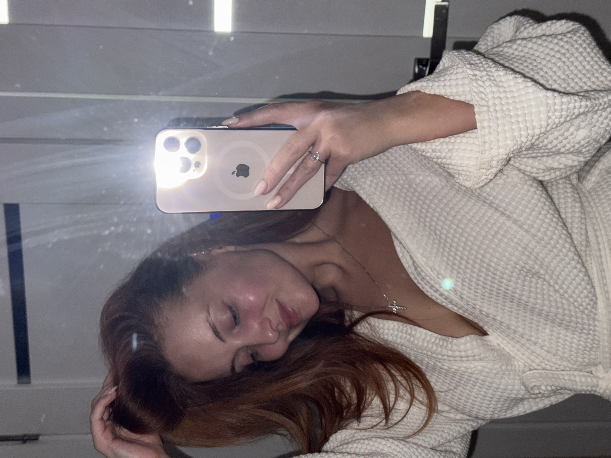 A woman is taking a selfie in front of a mirror. She is wearing a white waffle-weave bathrobe. Her hair is reddish-brown and partially covers her face. She is holding a smartphone with its flash activated. The phone has a round Apple logo and three camera lenses. The reflection shows a wall with horizontal and vertical white fixtures. The location is unknown and therefore cannot be confirmed to be Chaikovsky, Russia.