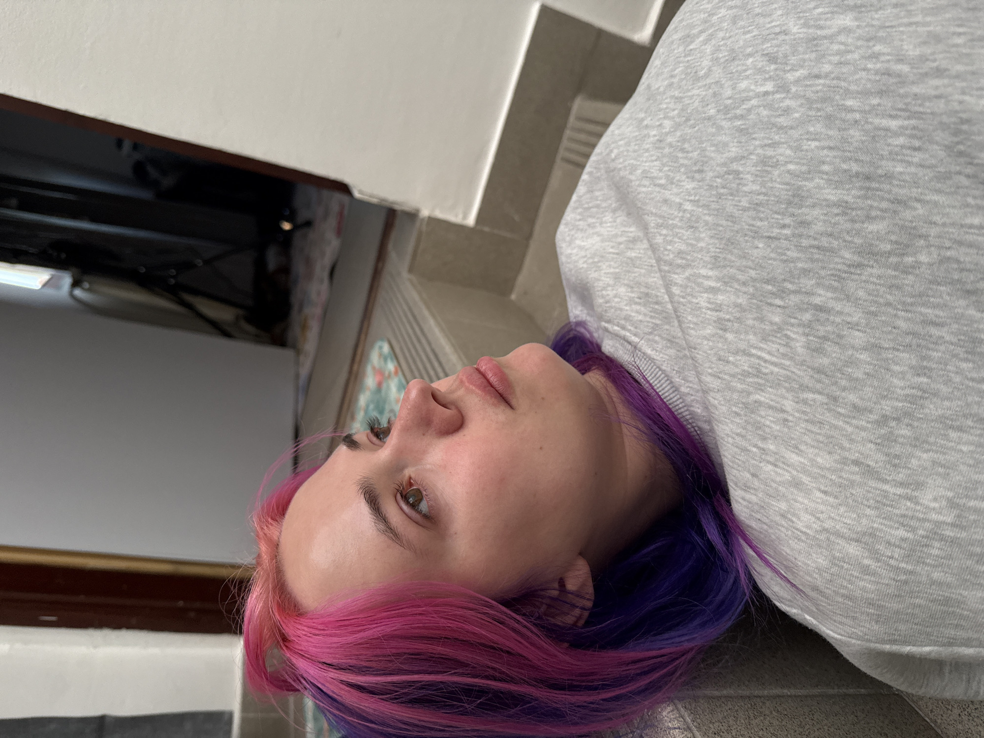 A young woman with hair dyed pink and purple is lying on a tiled floor near a set of stairs and a white wall or surface. She is wearing a gray sweatshirt. Based on the location information, the scene is in City of Zagreb, Croatia, but specific details about the location cannot be determined from the image.
