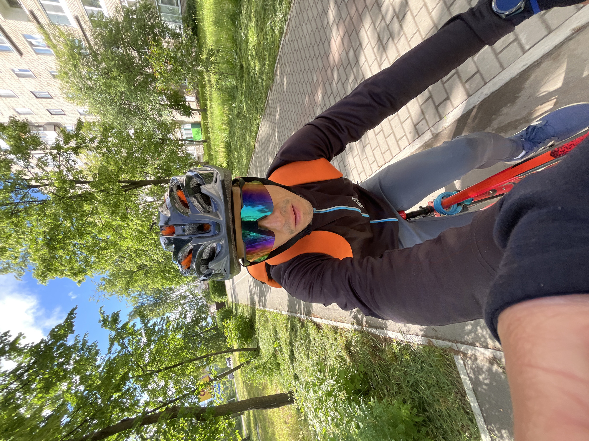 A person is seen in a selfie-style shot, seated on a red bicycle outdoors, likely cycling or paused during a ride. The individual, appearing male, wears a dark cycling helmet with orange accents and large, reflective, multi-colored sports sunglasses. They are dressed in a black long-sleeved cycling jersey or jacket with an orange collar and blue stripe, grey pants, and blue sneakers. A "NORDKAP" logo is partially visible on the jacket. A blue smartwatch is worn on the left wrist.
The setting appears to be an urban or suburban area in Chaikovsky, Russia, on a sunny day with a clear blue sky and some white clouds. Lush green trees line a paved sidewalk or path where the person is. In the background, a multi-story brick building with numerous windows is visible, suggesting a residential complex. Shadows are cast on the paved ground, indicating bright daylight. The overall impression is that of an active outdoor scene on a pleasant day.