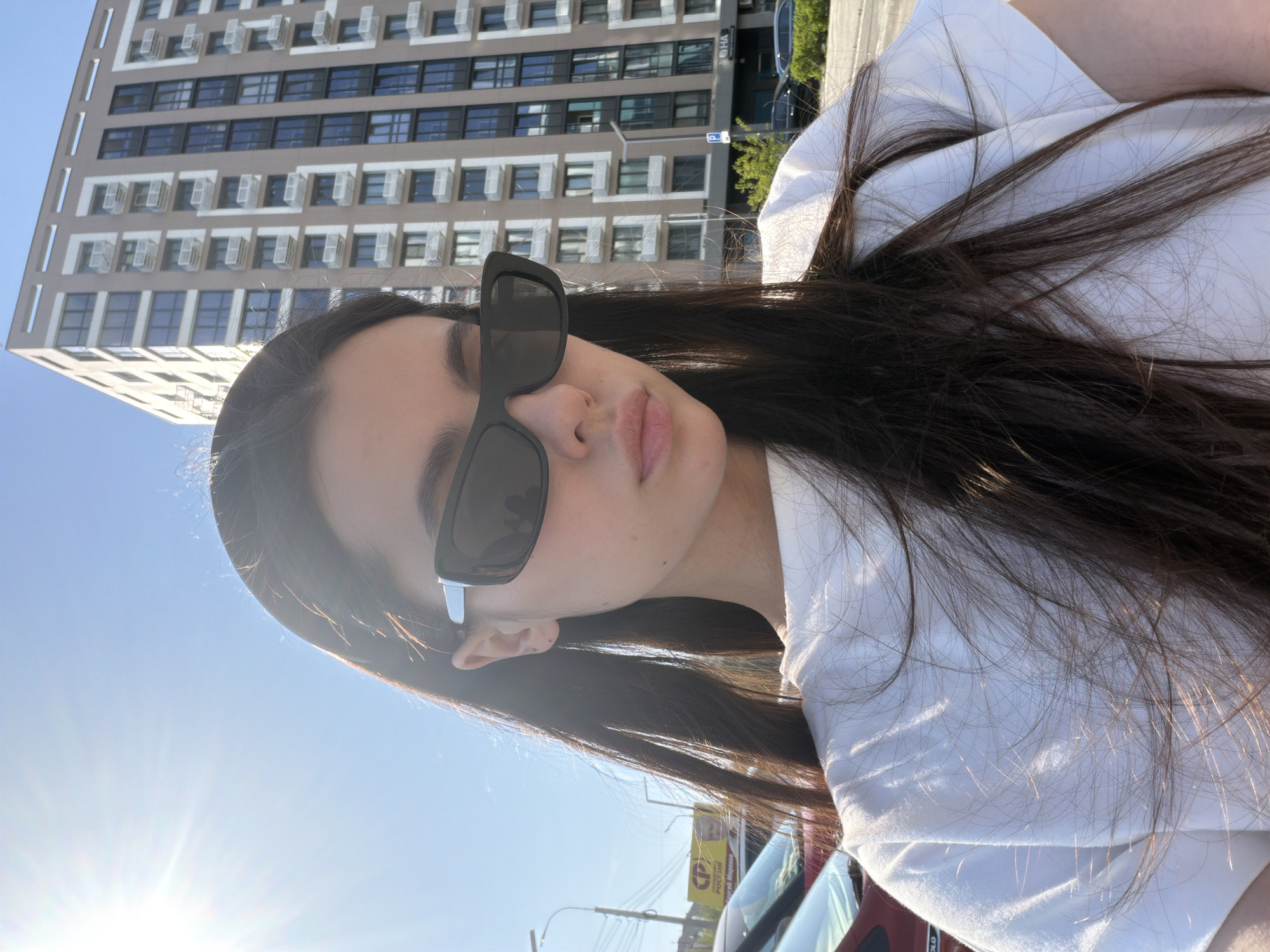 The image is an upward-angle selfie of a woman with dark hair wearing dark sunglasses and a white top. She is outdoors, likely in an urban environment. In the background, there is a multi-story building with numerous windows, possibly an apartment building. Streetlights and possibly traffic can be seen at the bottom of the frame. Based on visual elements (e.g., Cyrillic script on a pole and architectural style), the location may be the city of Izhevsk, Russia. The photograph appears to have been taken during the daytime.