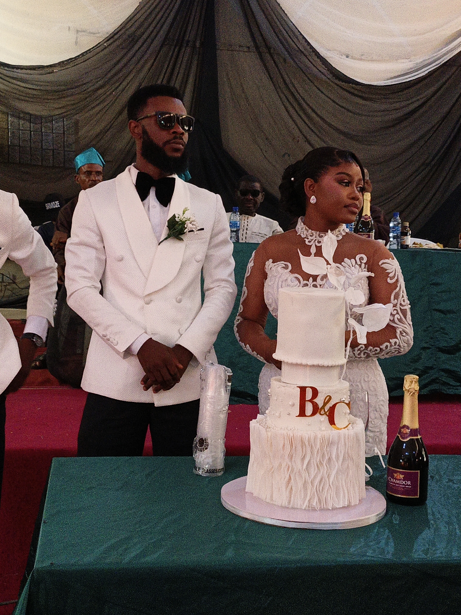 In Fagge D2, Nigeria, a wedding scene unfolds indoors, evidenced by draped black and white fabric in the background. A man in a white double-breasted suit, black bow tie, and sunglasses stands to the left of a three-tiered white wedding cake adorned with the initials "B&C" in metallic lettering. A woman in a white lace dress with long sleeves and a high neckline stands to the right of the cake. A bottle of "Chamdor" non-alcoholic sparkling juice sits on the green tablecloth. In the background, several people are seated at a table. Another man in a white suit stands partially in the left foreground. Plastic cups and other objects are also on the table. The floor appears to be covered with red carpet.