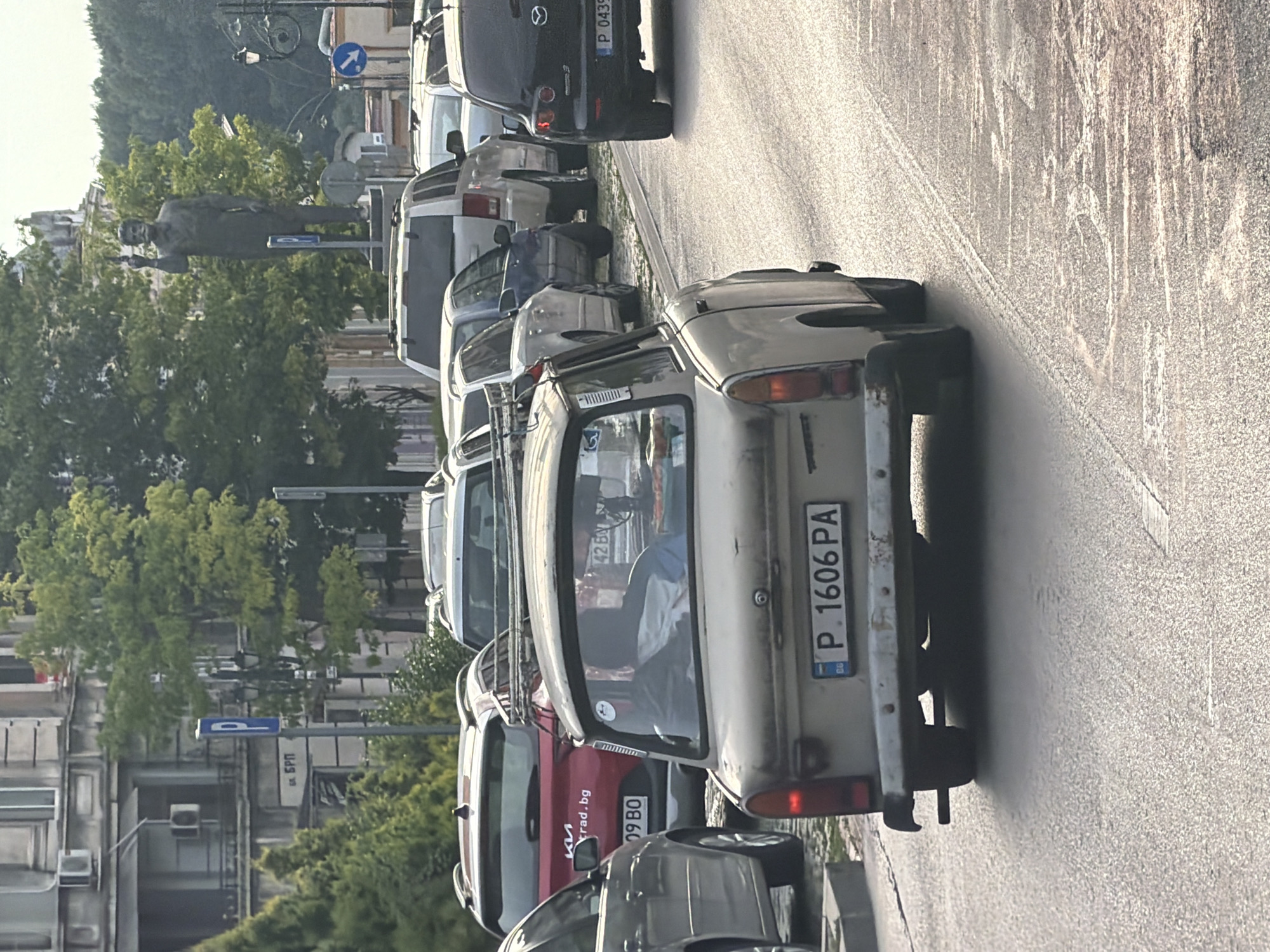 The image shows a street scene in Ruse, Bulgaria, with several cars parked and driving. The central focus is a light-colored, older model car, possibly a Trabant, with a license plate reading "P. 1606 PA". This car is in motion. In the background, other vehicles, including a red SUV with a "rad.bg" sticker and a black Mazda, are visible. A statue of a man is prominent in the distance, along with buildings and trees, suggesting an urban environment. Parking signs are also present. The lighting indicates it is daytime, likely late afternoon given the angle of the shadows.