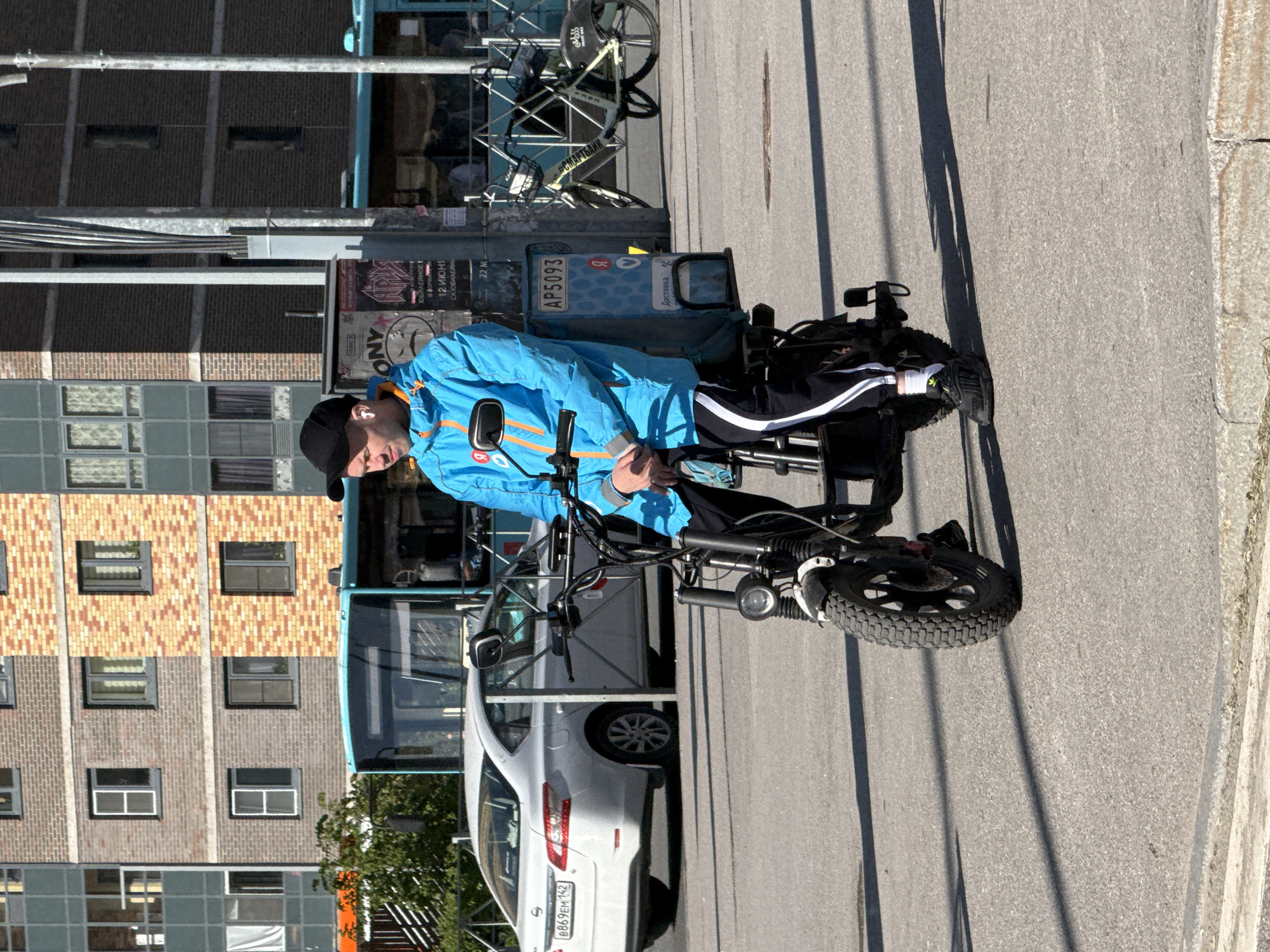 The image captures a man stopped on an electric scooter on a sunny day in an urban environment. The man, seen from the waist up, wears a bright blue jacket with orange accents, a black baseball cap, and white earbuds. He is seated on a black electric scooter or moped-style bike with large, knobby tires, and appears to be looking down at something in his hands. His lower body is clad in black track pants with white stripes down the sides and black sneakers with white socks.
A blue delivery box is attached to the rear of his scooter, featuring the number "AP5093," the Russian letter "Я" (likely part of a company logo like Yandex), and the word "Доставка" (Delivery). This suggests the man is a delivery rider.
In the background, a light-colored sedan, possibly a Nissan, is parked, displaying a Russian license plate "В869ЕМ 192." A blue city bus is partially visible behind the car. Further back, multi-story buildings with brick and concrete facades line the street, featuring numerous windows.
Near the bus, a bicycle rack holds several bicycles, one of which has a sign with "#СМАРТБЕЙК" (#Smartbike). On a pole next to the bus, various posters or signs are attached, with visible Russian text including "ИЮНЯ" (June), "В ФОРМЕ" (In shape), "СМАРТЕБАН," and a partial "ONY." The bright sun casts distinct shadows on the asphalt, indicating it is daytime with clear weather.