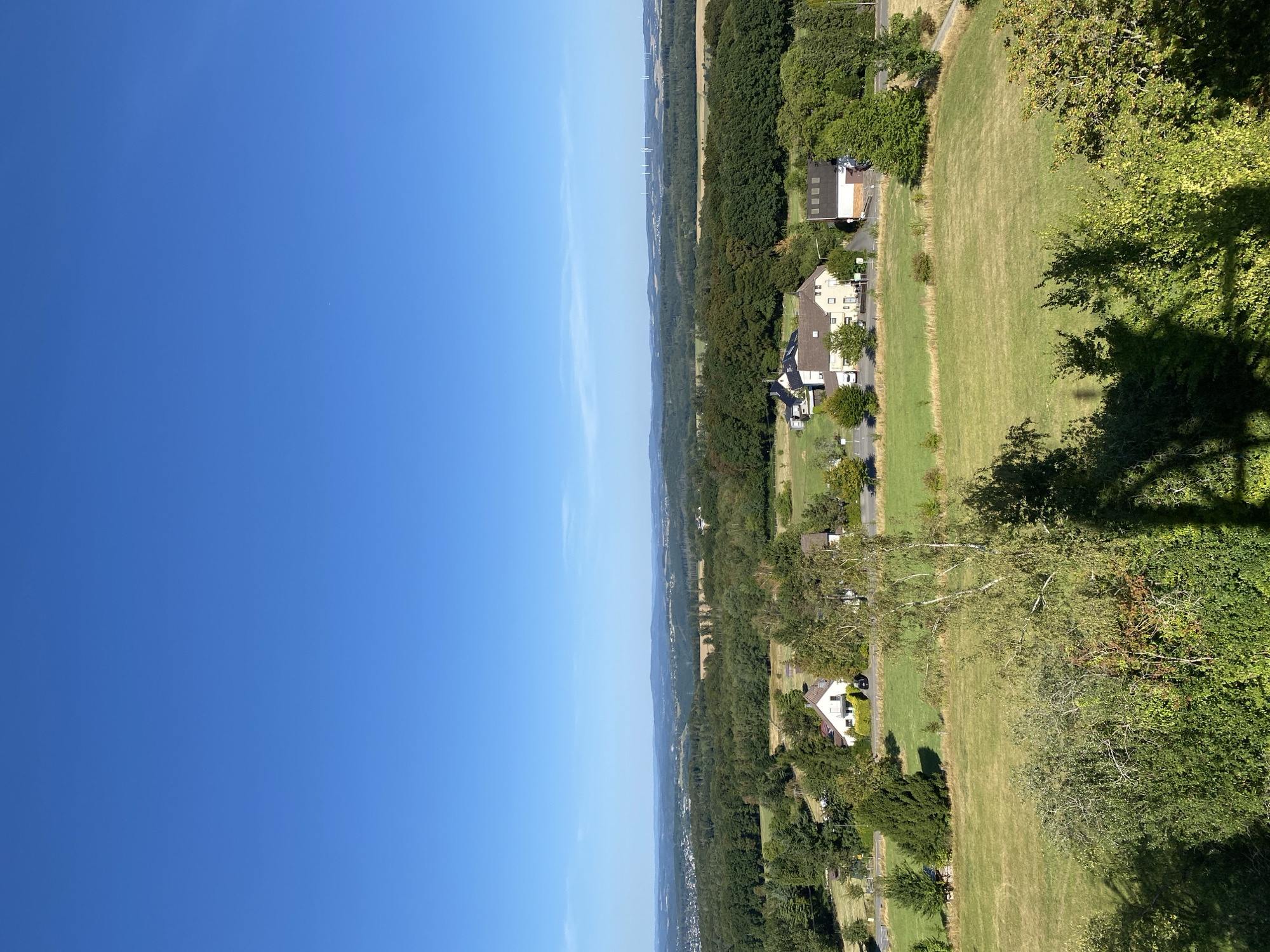 An aerial, high-angle, full shot captures a panoramic view of a rural landscape under a clear blue sky in Busenhausen, Germany. The image is dominated by a vast expanse of greenery, with rolling hills and dense forests stretching into the distance. In the mid-ground, several residential buildings are nestled among the trees, characterized by their white and beige facades and pitched roofs. A paved road winds through the scene, connecting the houses and leading to a parking area. The foreground features lush green fields and trees, with a visible shadow cast on the foliage, suggesting the presence of an unseen object or structure from which the photograph was taken. The distant horizon is marked by a chain of hills and several wind turbines, indicating a relatively open and developed area despite the rural setting. The overall atmosphere is serene and idyllic, bathed in bright daylight.