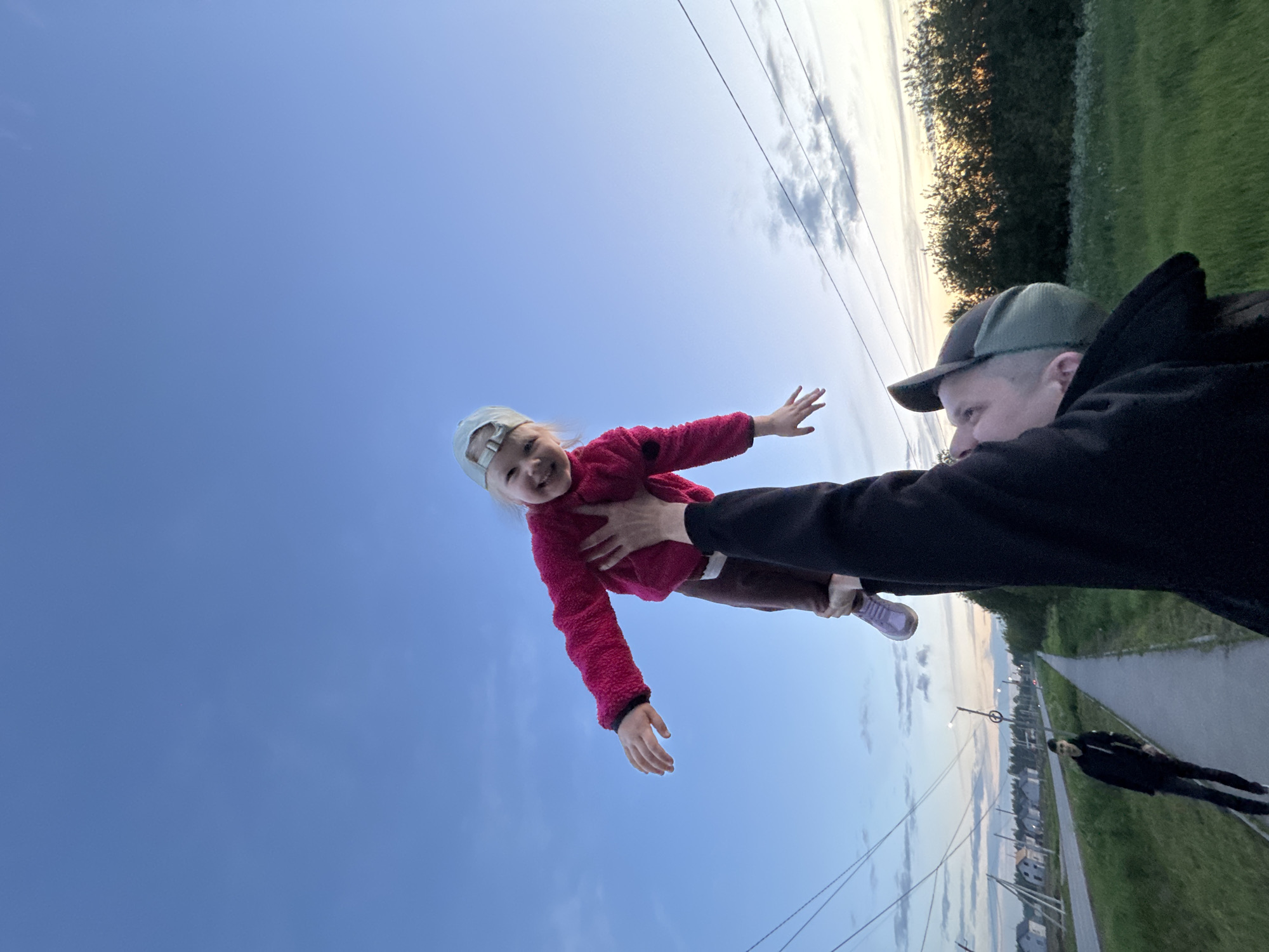A man is holding a young girl up in the air. The girl is wearing a red jacket, brown pants, and a baseball cap. The man is wearing a black hoodie and a green baseball cap. The sky is blue and there are power lines visible. In the background, there is a path and a person walking on the path. The location appears to be a city neighborhood in Горный, Russia.