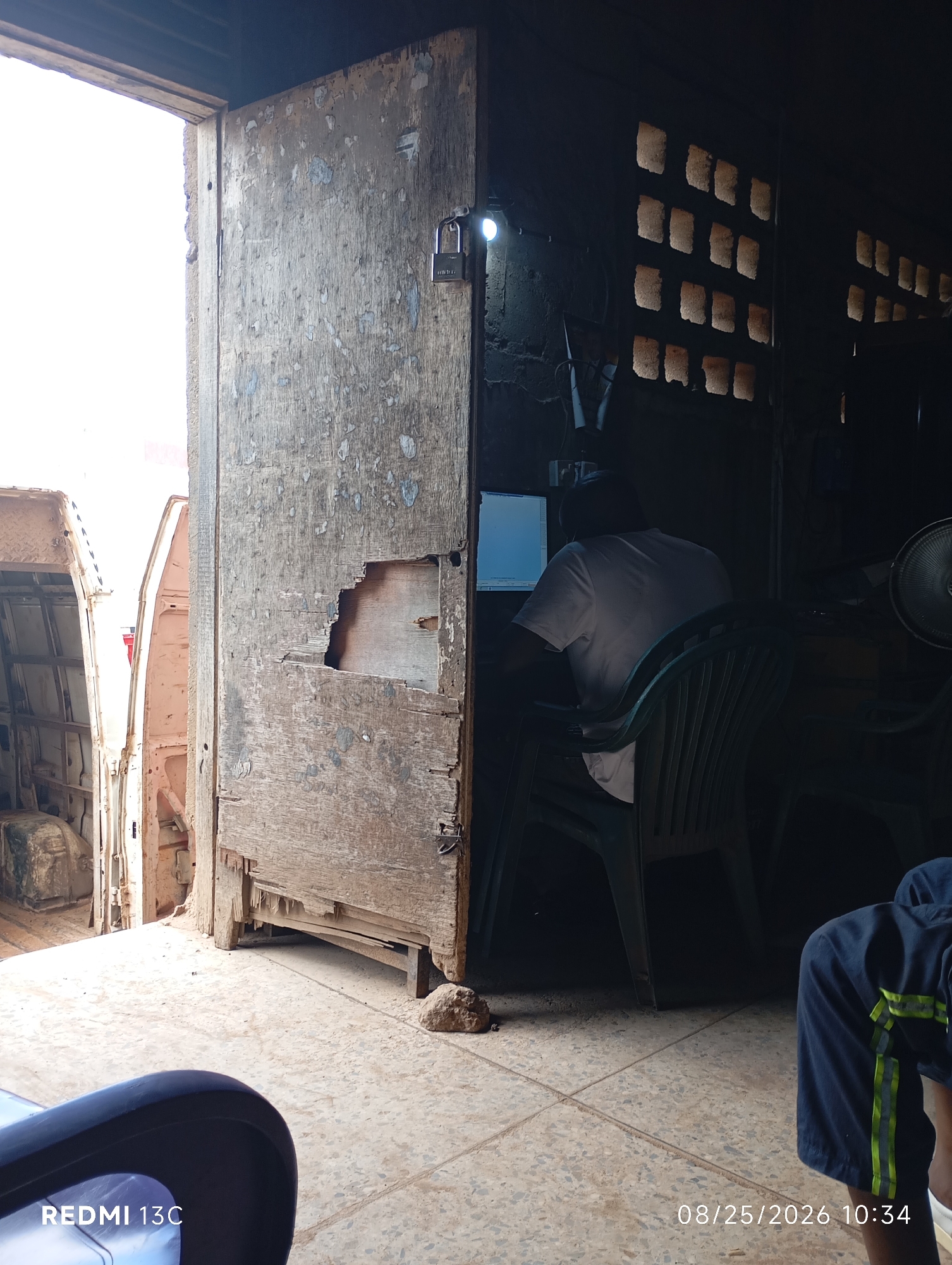 The image captures an interior scene, likely a shop or workshop, in Shendam, Nigeria. A heavily weathered and damaged wooden door is a prominent feature, standing ajar to reveal a bright outdoor environment. The door has a padlock attached to it. Inside, the space is dimly lit. A person in a white t-shirt is seated at a desk, facing a computer monitor. Two green plastic chairs are visible, one occupied by the person at the computer and another empty chair. In the background, a wall with a series of square openings, possibly ventilation or windows, is visible, with light filtering through. To the left of the door, the rear section of a white vehicle, possibly a van or truck, is visible, suggesting the shop may be an auto-related business or located near one. The floor is tiled. A portion of the lower right of the image shows another person's legs wearing dark shorts with neon green stripes and a white shoe. The date and time stamp "08/25/2026 10:34" and "REDMI 13C" are overlaid on the bottom of the image.