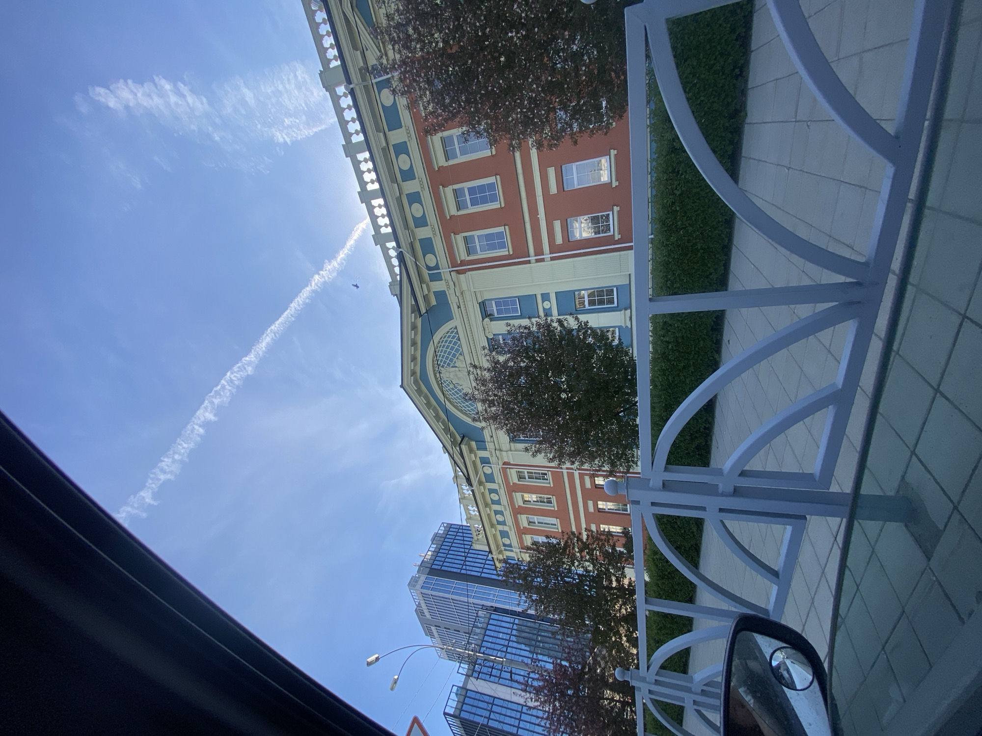 The image shows a view from inside a vehicle, likely a car, looking out towards the architecture of Yekaterinburg, Russia. A white, ornamental fence runs along the right side of the frame, partially obstructing the view.
Buildings visible include a red and blue building with multiple windows, reminiscent of a classical or historical style, and a modern, glass-covered skyscraper. The buildings are framed by trees with reddish leaves.
A contrail is visible in the blue sky. A car side mirror is partly visible in the bottom right corner. The interior of the vehicle, including the roof lining, is visible on the left side of the frame.