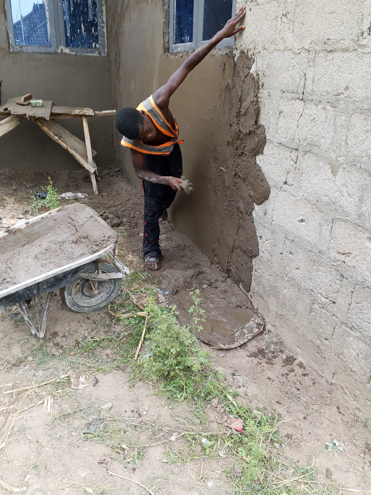 An outdoor scene in Adonkolo, Nigeria, depicts a construction worker actively plastering a building wall during the daytime. The main subject is a dark-skinned male, likely an adult, with short dark hair. He is wearing a sleeveless orange high-visibility vest over a dark top, dark trousers with red patterns, and sandals.
The worker is captured mid-action, leaning towards a concrete block wall to apply wet cement or plaster. His right arm is extended upwards, with his hand resting on the plastered section of the wall, possibly for balance or to gauge the finish. His left hand, blurred by movement, is near the wall, likely in the process of applying material with a tool. A significant portion of the wall to his left is already covered with a rough layer of dark gray plaster, while the section he is working on shows the wet, freshly applied material over the lighter, unfinished concrete blocks.
To the left of the worker, a black wheelbarrow is visible in the foreground, filled with a substantial amount of wet cement or plaster mix. Further back, against the wall, is a rudimentary wooden stand or table holding a couple of sponges or cleaning pads, also covered in the construction material. The wall itself features two windows with dark, reflective panes, suggesting they are already installed.
The ground around the work area is uneven, a mixture of dry earth, patches of green weeds and grass, and areas of wet mud, particularly where plaster has fallen or been spilled. A discarded, muddy sheet or bag lies on the ground near the wall. The overall setting is a functional, active construction site, characterized by manual labor and basic building materials. No visible text is present in the image.