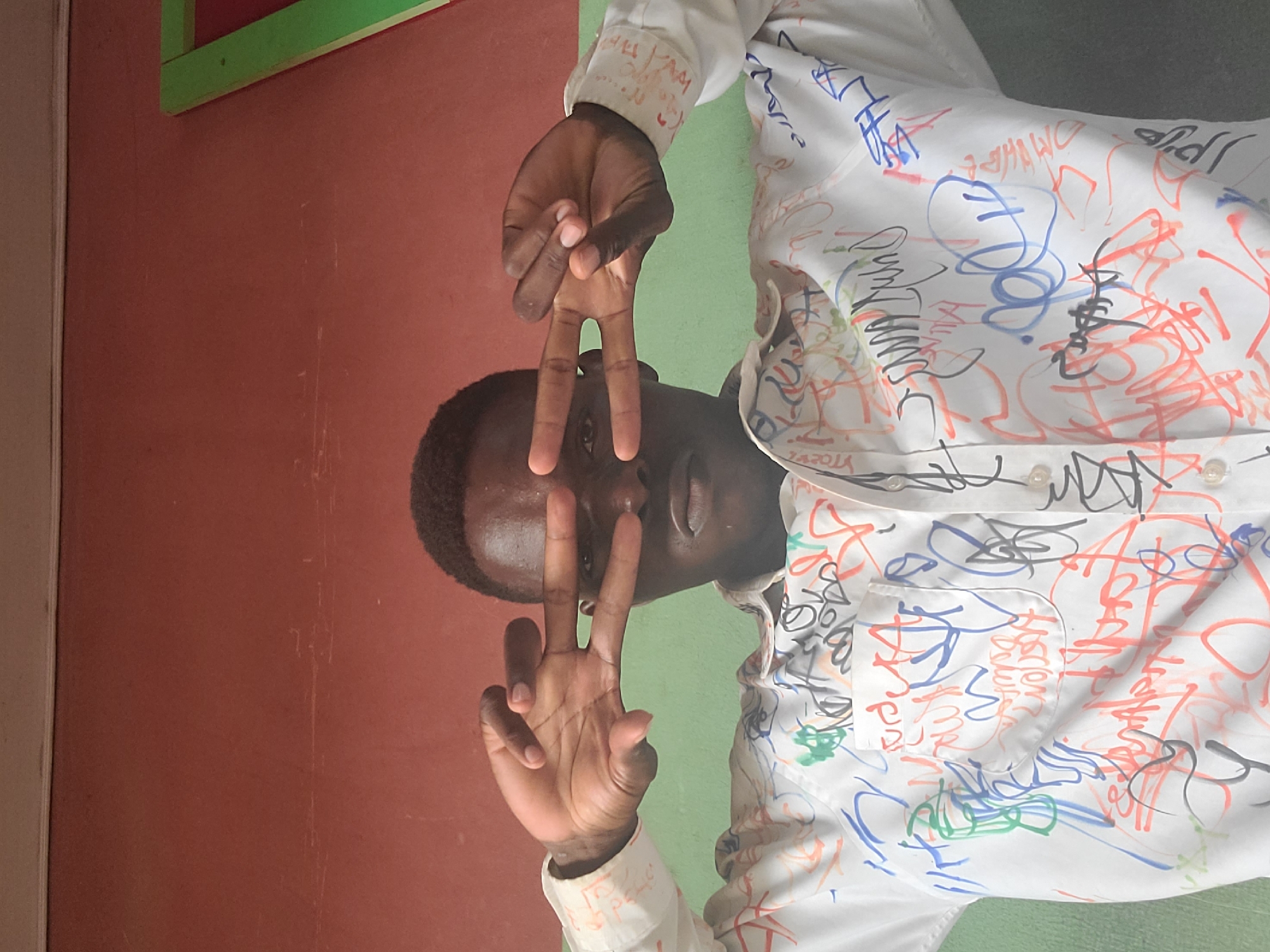 The image shows a close-up of a young man's face and upper torso, taken indoors against a backdrop of a reddish-brown wall and a green section of a wall or door frame. The man is wearing a white button-down shirt covered in colorful scribbled drawings and graffiti. He is looking directly at the camera, and his hands are held up in front of his face, forming a "peace" sign with his right hand and framing his eyes with his fingers. The lighting suggests an indoor setting, possibly natural light. The environment appears to be a room with simple walls, and the overall impression is casual and informal. There are no visible indications of the specific location within Gura Top, Nigeria, beyond the general context of an indoor setting with painted walls.