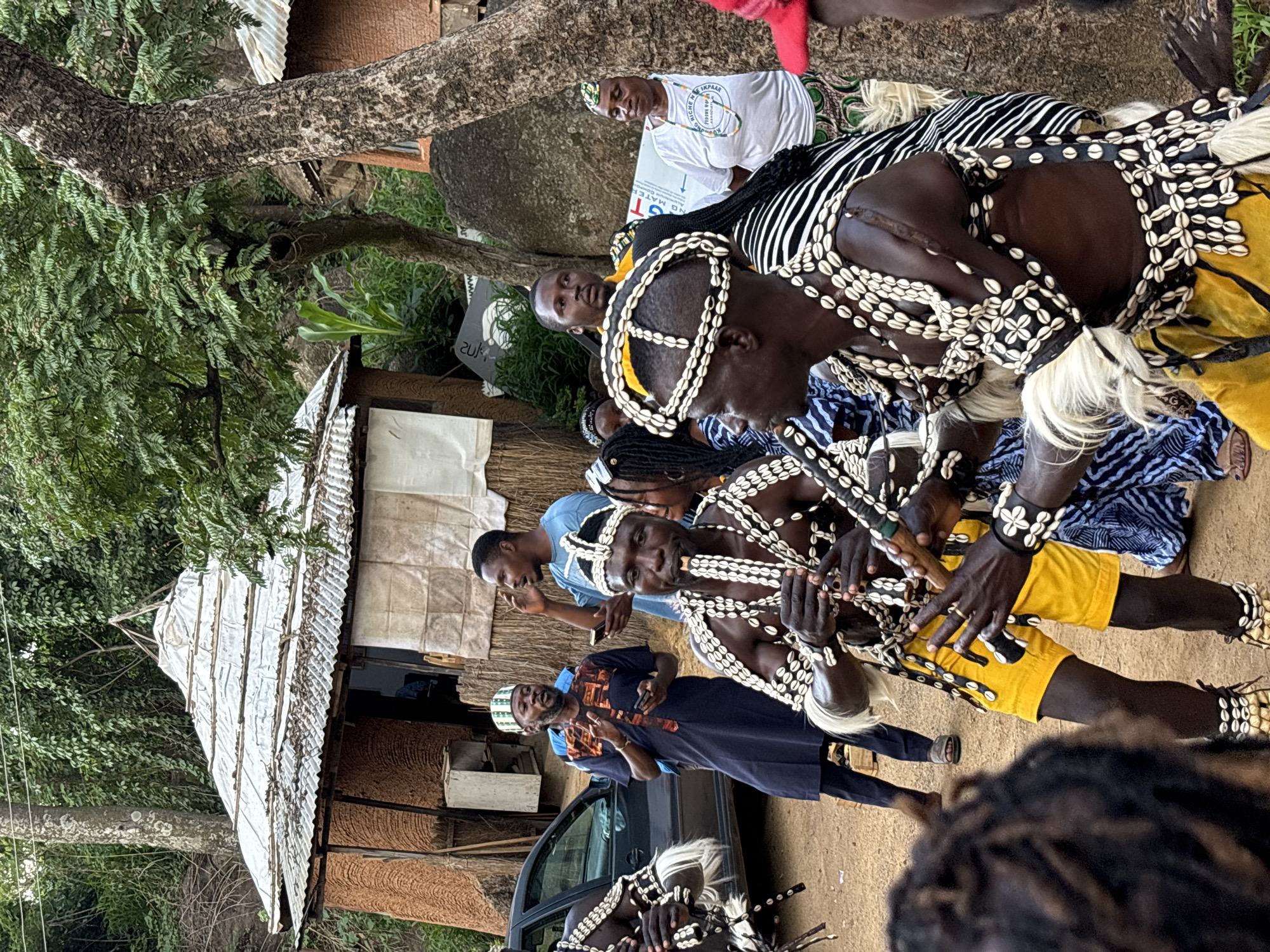 A group of people are gathered in an outdoor, dusty area with some traditional huts in the background. The scene appears to be in a rural or semi-rural setting, possibly in Jos, Nigeria. In the foreground, two men are dressed in elaborate traditional attire adorned with numerous cowrie shells and feathers. One of them is playing a flute-like instrument, while the other is also holding a similar instrument. Their costumes include headdresses, chest ornaments, armbands, and leg bands, all decorated with shells.
Several other individuals are present, observing the performers. One man in a dark blue and orange patterned robe is speaking with his hands raised, holding a mobile phone. To his right, another man in a blue t-shirt is talking, also holding a phone. Further back, a woman in a white t-shirt with text and graphics is looking towards the performers, and another woman in a colorful patterned dress is partially visible. A dark-colored car is parked to the left of the scene.
The environment includes dense green foliage, trees with textured bark, and large rocks. The huts in the background are constructed with natural materials, featuring thatched or corrugated metal roofs. The ground is covered in dirt and sand. The overall atmosphere suggests a cultural event or performance.