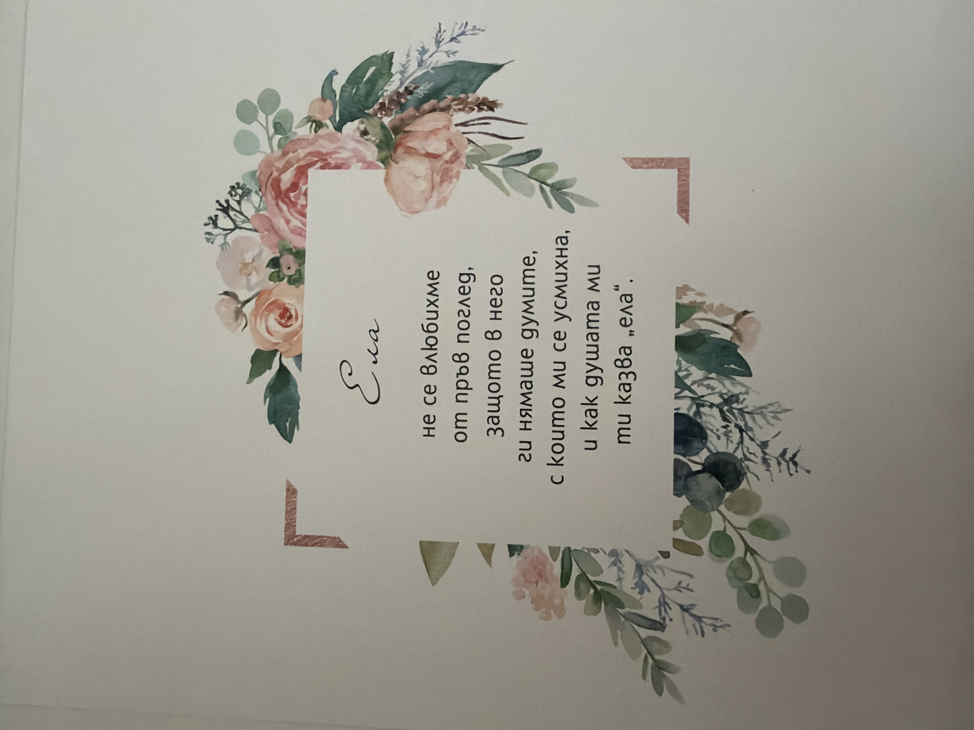 The image shows a greeting card with a floral design and text in Bulgarian. The text appears to be a poem or a message. The floral design features roses and other greenery in soft, watercolor tones, framing a white rectangular area containing the text. The background of the card is a light, textured surface, likely paper. There are no people or animals present in the image. The text reads: "Ела не се влюбихме от пръв поглед, защото в него ги нямаше думите, с които ми се усмихна, u kak душата mu tuказва „ела“." This translates to: "Come, we did not fall in love at first sight, because in it there were no words, with which you smiled at me, and how your soul tells you "come"."
There is no information in the image that would indicate its location in the city of Pazardzhik, Bulgaria.