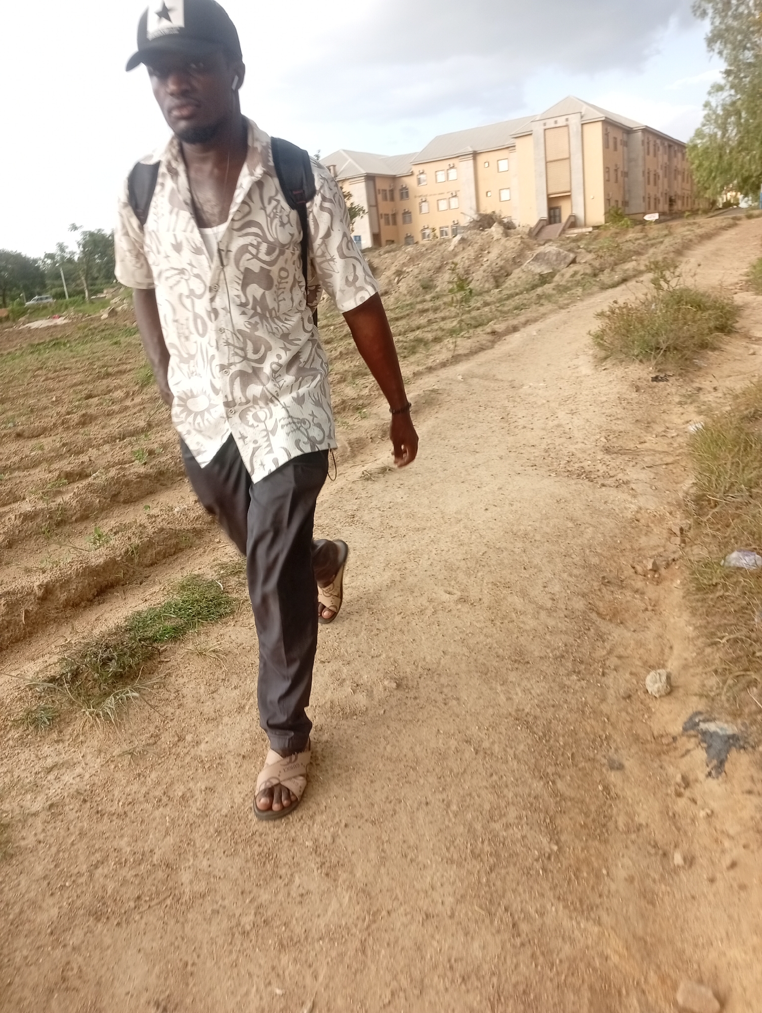 A young Black man wearing a black baseball cap with a white star, a patterned short-sleeved shirt over a white undershirt, dark pants, and light brown sandals is walking on a dirt path in an outdoor, somewhat overgrown environment. He has a backpack on his back, and he is looking down. In the background, there is a multi-story tan building with many windows, and a dirt embankment leading up to it. The sky is overcast. The area appears to be on the outskirts of Jos, Nigeria, possibly a developing area or a construction site, with evidence of terraformed land and sparse vegetation.