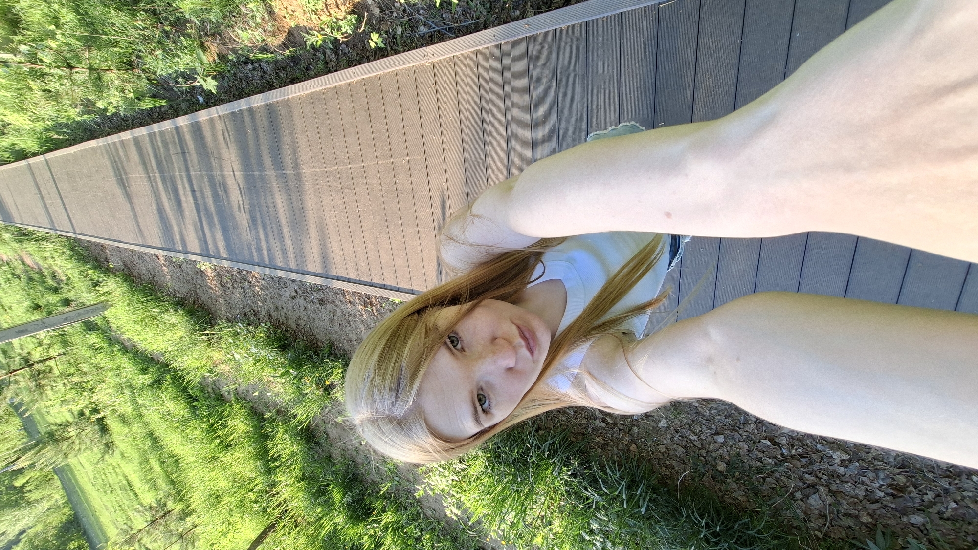 A woman with blonde hair and a white tank top is taking a selfie on a wooden walkway. To the left of the walkway is grass and trees. To the right is grass and gravel. The location appears to be in an outdoor area with natural elements. There are trees and a walking path. The image does not give enough information to identify the location as the city of Chaikovsky, Russia.