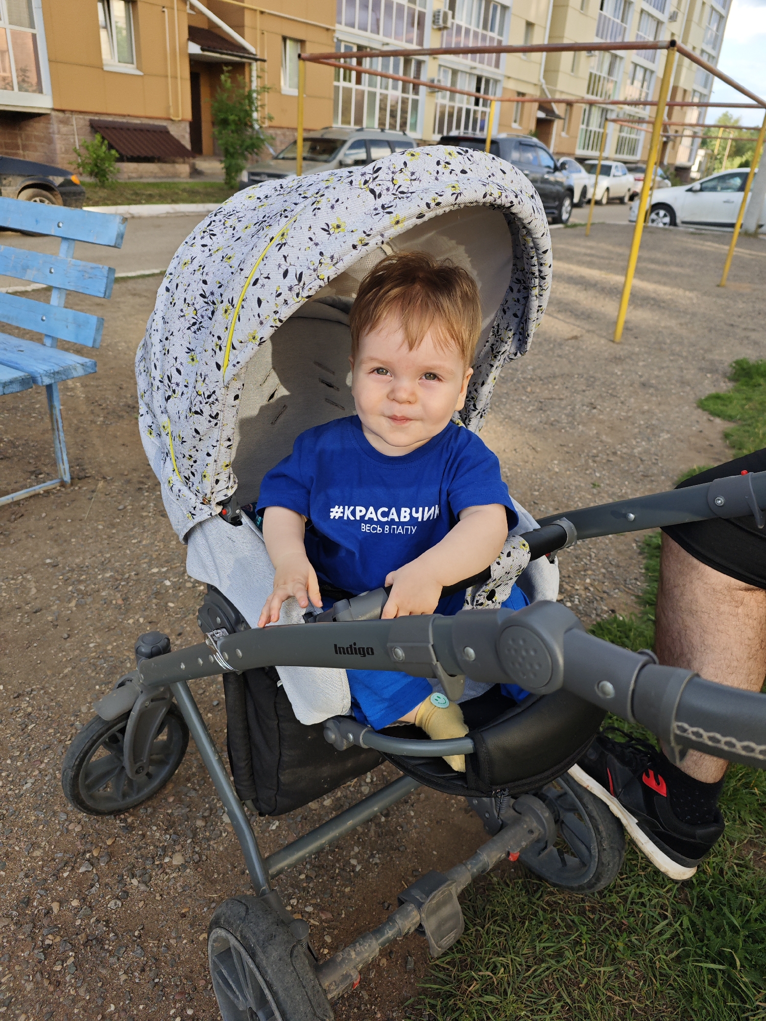 An outdoor shot captures a young child sitting in a stroller. The child, with light brown hair and fair skin, is wearing a blue t-shirt with white text. The stroller has a grey frame and a patterned canopy. The background features a building with yellow siding, a blue bench, and parked cars. The ground is a mix of dirt and gravel. The overall impression is a casual moment in a residential area.