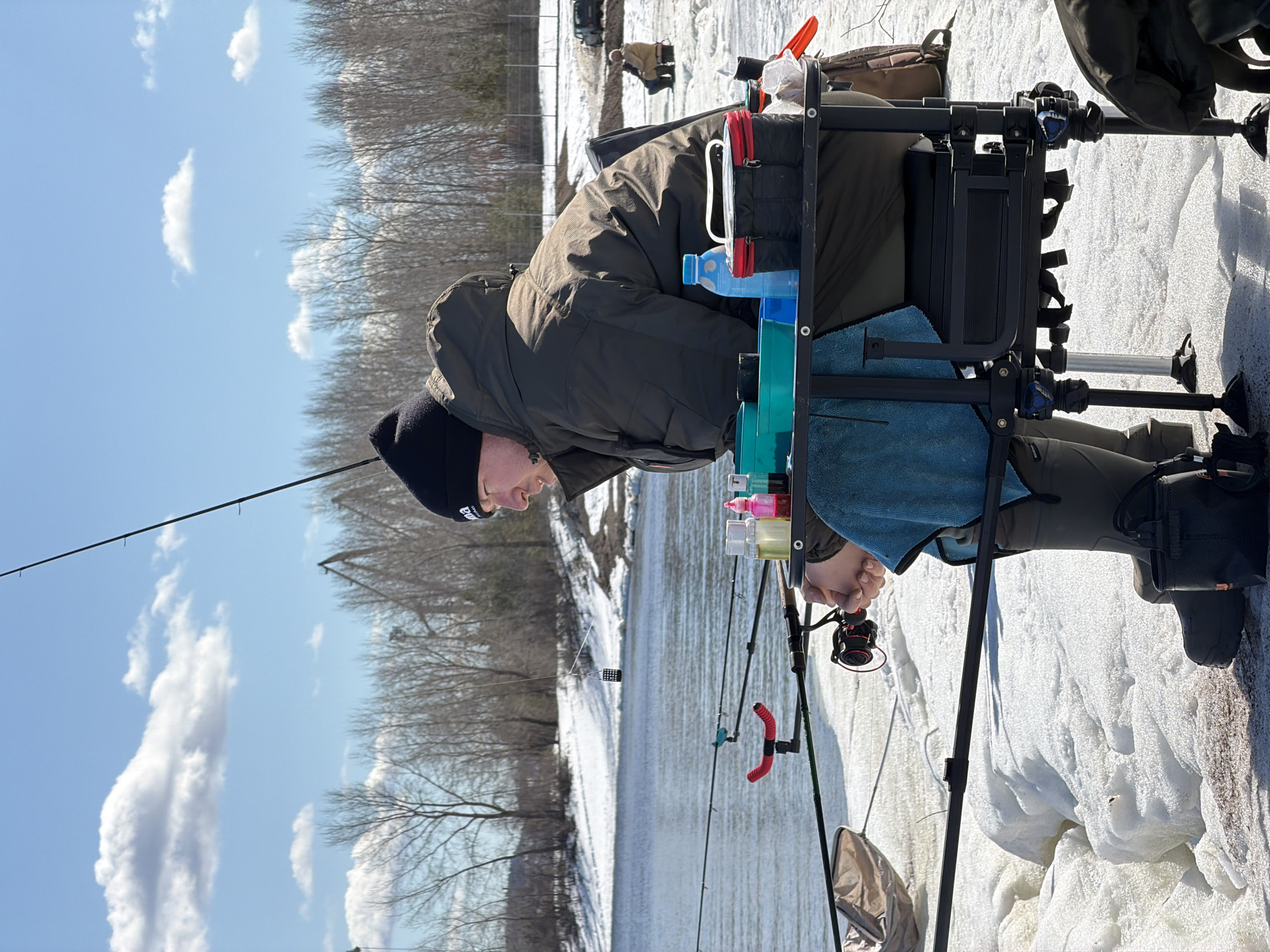 A man in a dark winter coat and black hat is ice fishing. He sits on an elevated fishing chair or platform, surrounded by fishing gear, including multiple rods and bait containers. The ground is covered in snow and ice, indicating a cold climate. The sky is partly cloudy. In the background, bare trees line the edge of a field. Another person is further in the background, also ice fishing. The location is suggested as Noviy, Russia, due to context. The man is holding a fishing rod and reel, actively fishing.