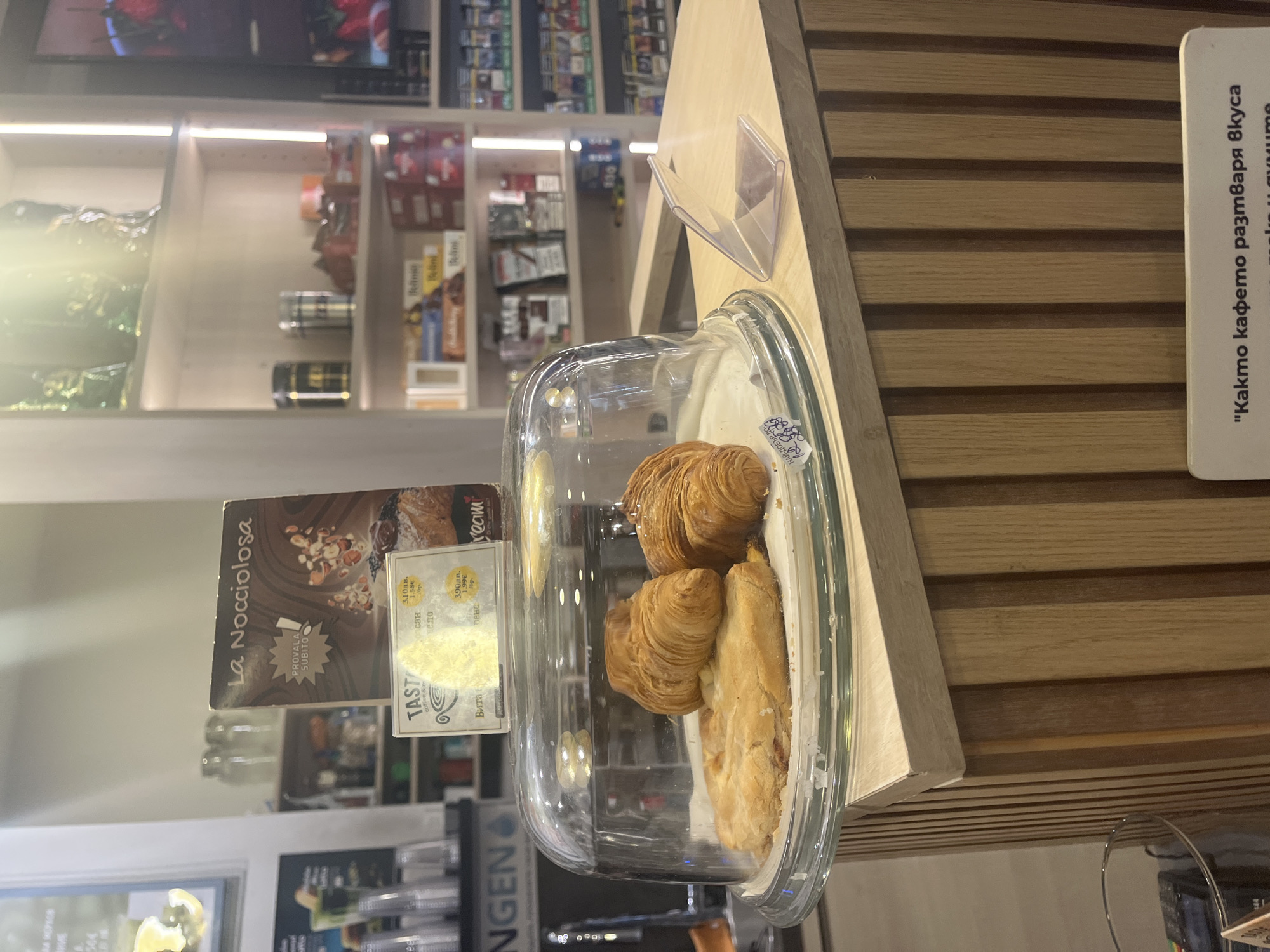 The image shows a pastry display in what appears to be a cafe or store in Plovdiv, Bulgaria. Under a clear dome are several croissants and a slice of pastry. A small sign is placed near the pastries, advertising "La Nocciolosa." A price tag is visible on the sign, listing prices in both Bulgarian Lev (лв) and Euros (€). In the background are shelves filled with various items, possibly including coffee, tea, and other food products. The setting is well-lit, suggesting an indoor commercial environment. A sign with Cyrillic text in a dark-wood, lined background reads: "Както кафето разтваря вкуса."