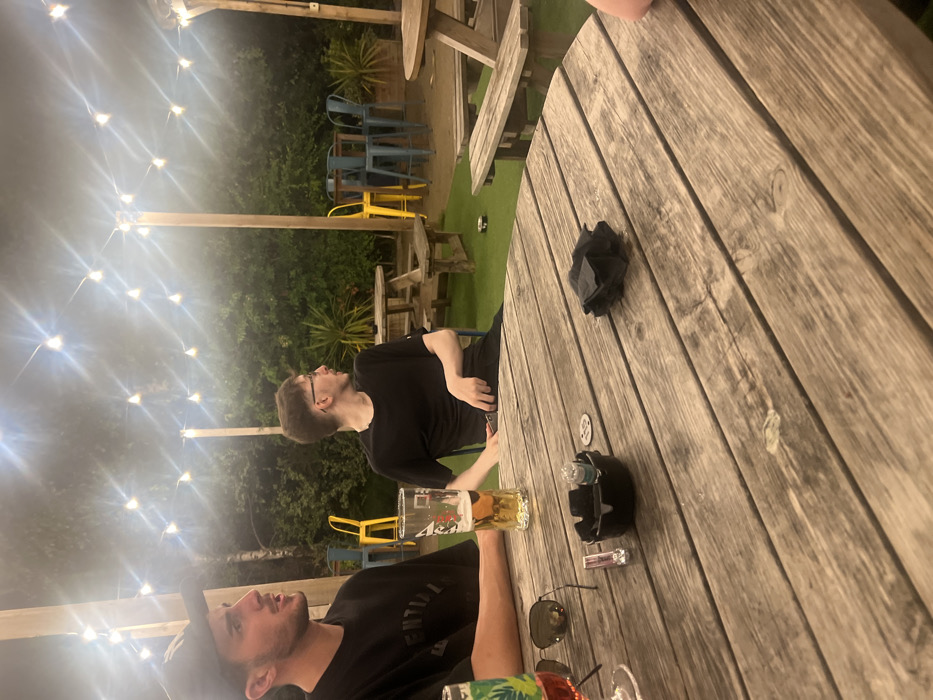 Two men sit at a wooden table under string lights. One man holds a beer, while the other looks off into the distance. An ashtray, sunglasses, lighter, and a crumpled napkin also sit on the table. A few tables and chairs are visible in the background, surrounded by plants and shrubbery.