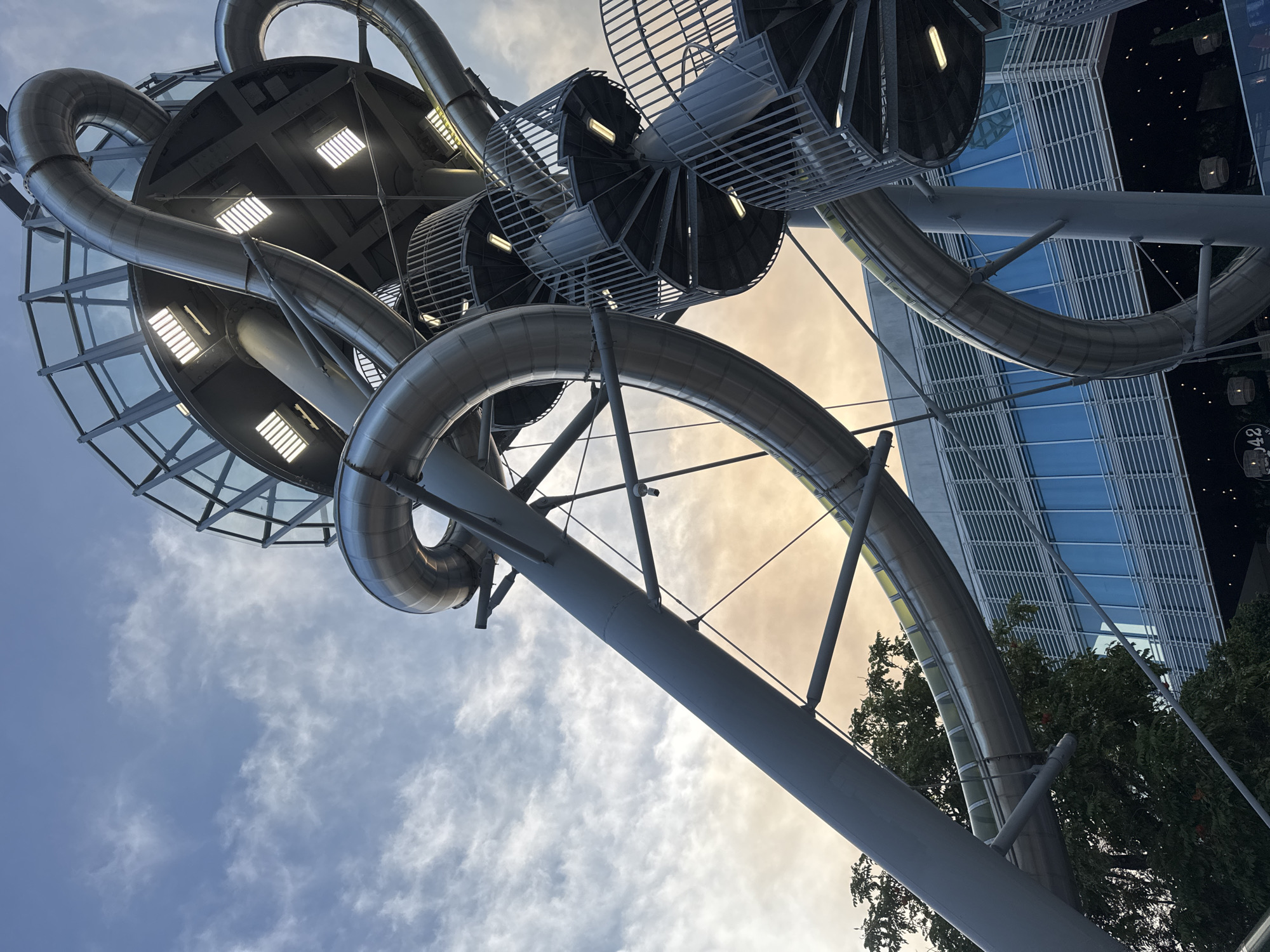 A low-angle outdoor shot captures a large, metallic slide structure against a cloudy sky at dusk. The slide, made of interconnected silver tubes, twists and turns upwards, forming a complex network. Several spiral staircases with metal railings ascend alongside the slides, leading to different levels of the structure. Lights are visible within some of the enclosed areas of the slide and on the staircases. In the background, a modern building with a glass facade and horizontal louvers is visible. Green trees are present at the bottom of the frame, and small lights, possibly from the building's facade or streetlights, are scattered at the very bottom. The sky displays a mix of blue and orange hues from the setting sun.